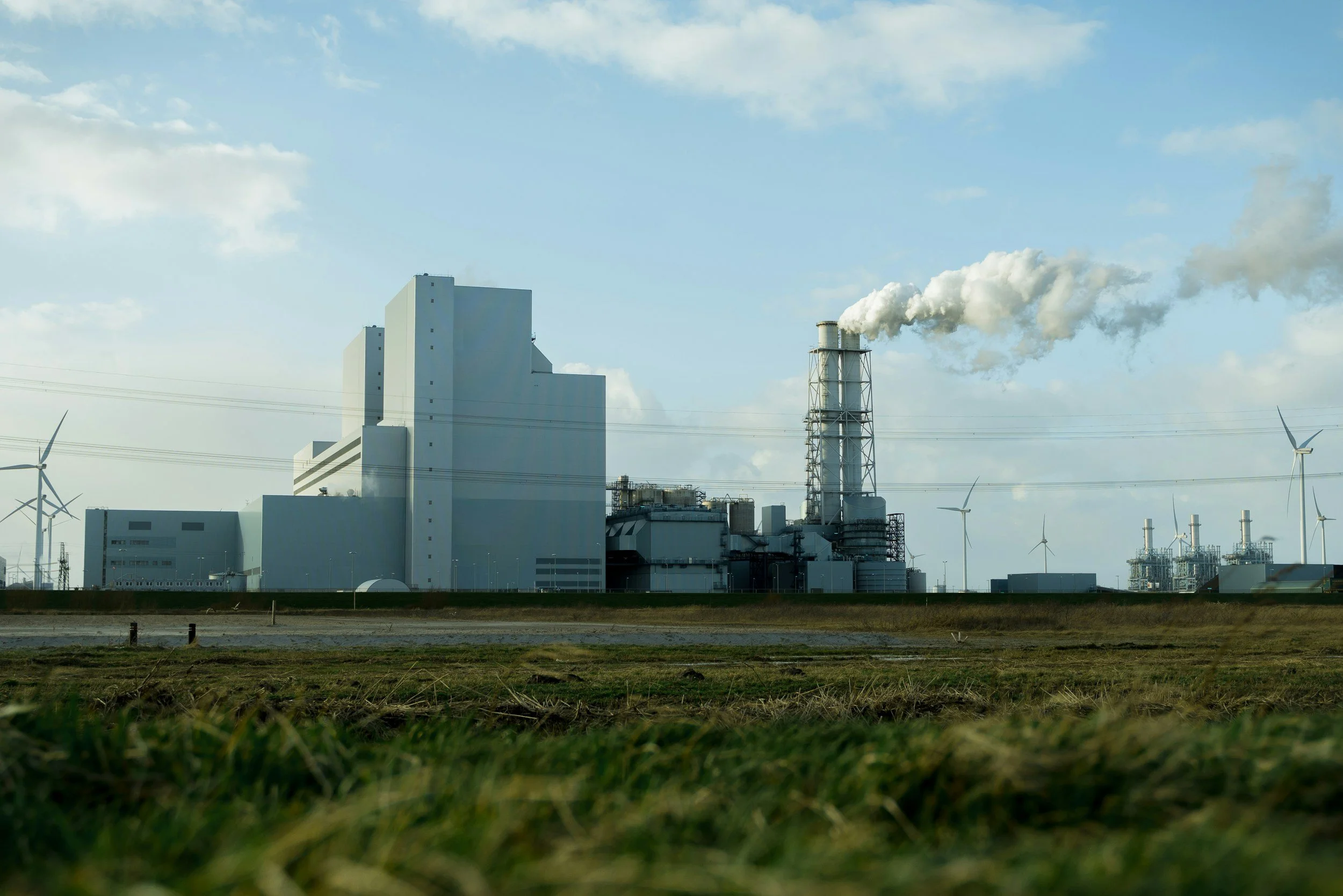 A coal power plant with large buildings, a smokestack emitting smoke, wind turbines in the background, and power lines in the foreground.