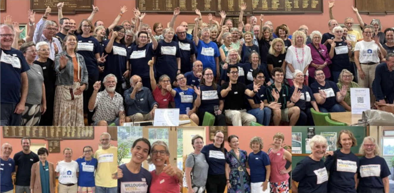 Group of people smiling and waving in a large room with a notice board and menu boards on the wall, some wearing matching shirts with printed messages, suggesting a voting or election event.