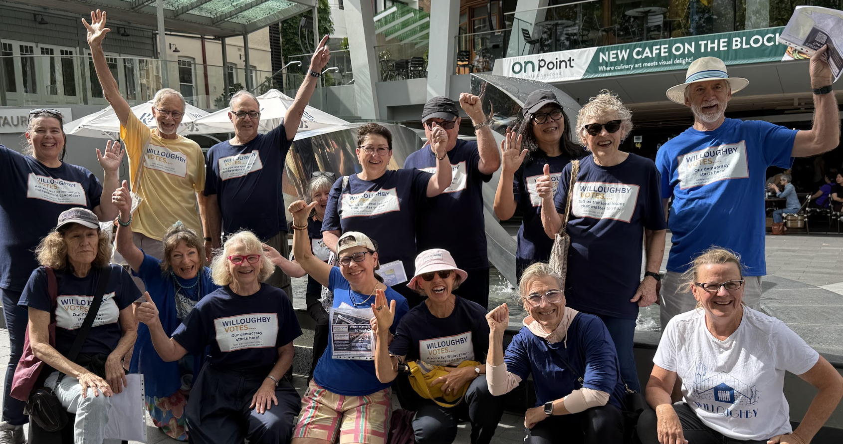 Group of diverse smiling people wearing 'Willoughby Votes' t-shirts, celebrating and raising their hands outdoors in front of modern buildings and cafes during daytime.