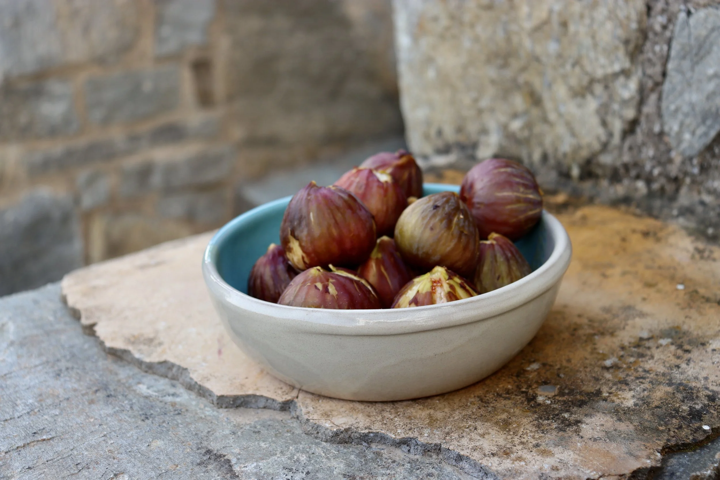 Figues dans un bol blanc avec intérieur turquoise, posé sur une surface en pierre, avec un mur en pierre en arrière-plan.