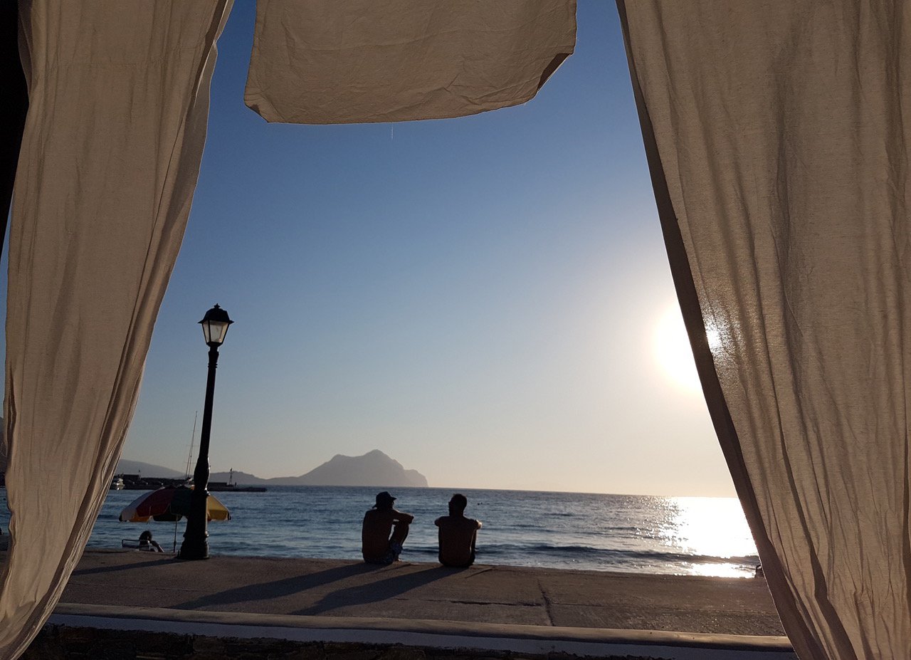 Deux personnes assises au bord de la mer, vues à travers une ouverture de tissu, avec un lampadaire, un parasol, et une montagne au loin, au coucher du soleil.