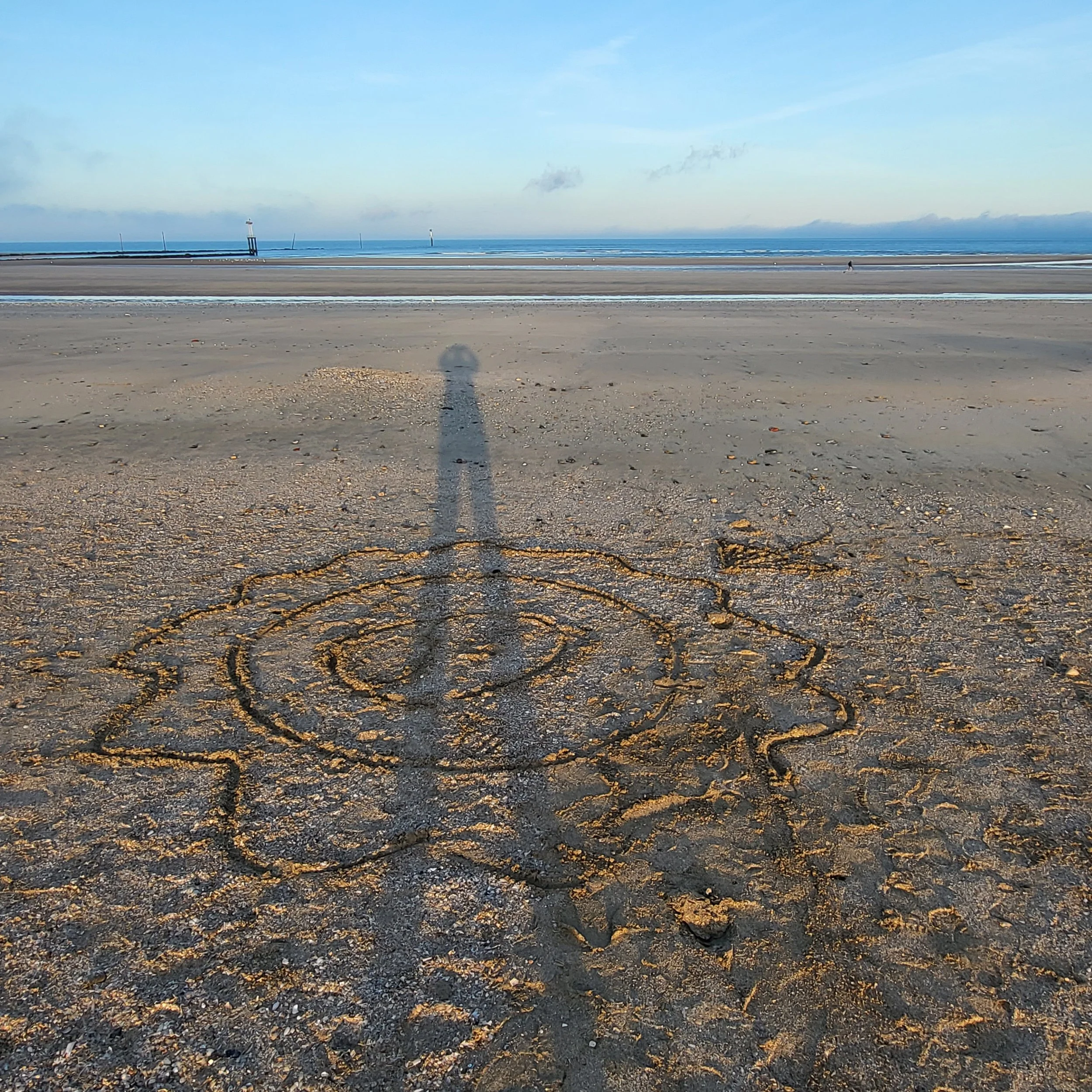 Ombre d'une personne sur une plage, avec un dessin en forme de papillon ou d'ange dans le sable en premier plan, et la mer à l'arrière-plan.