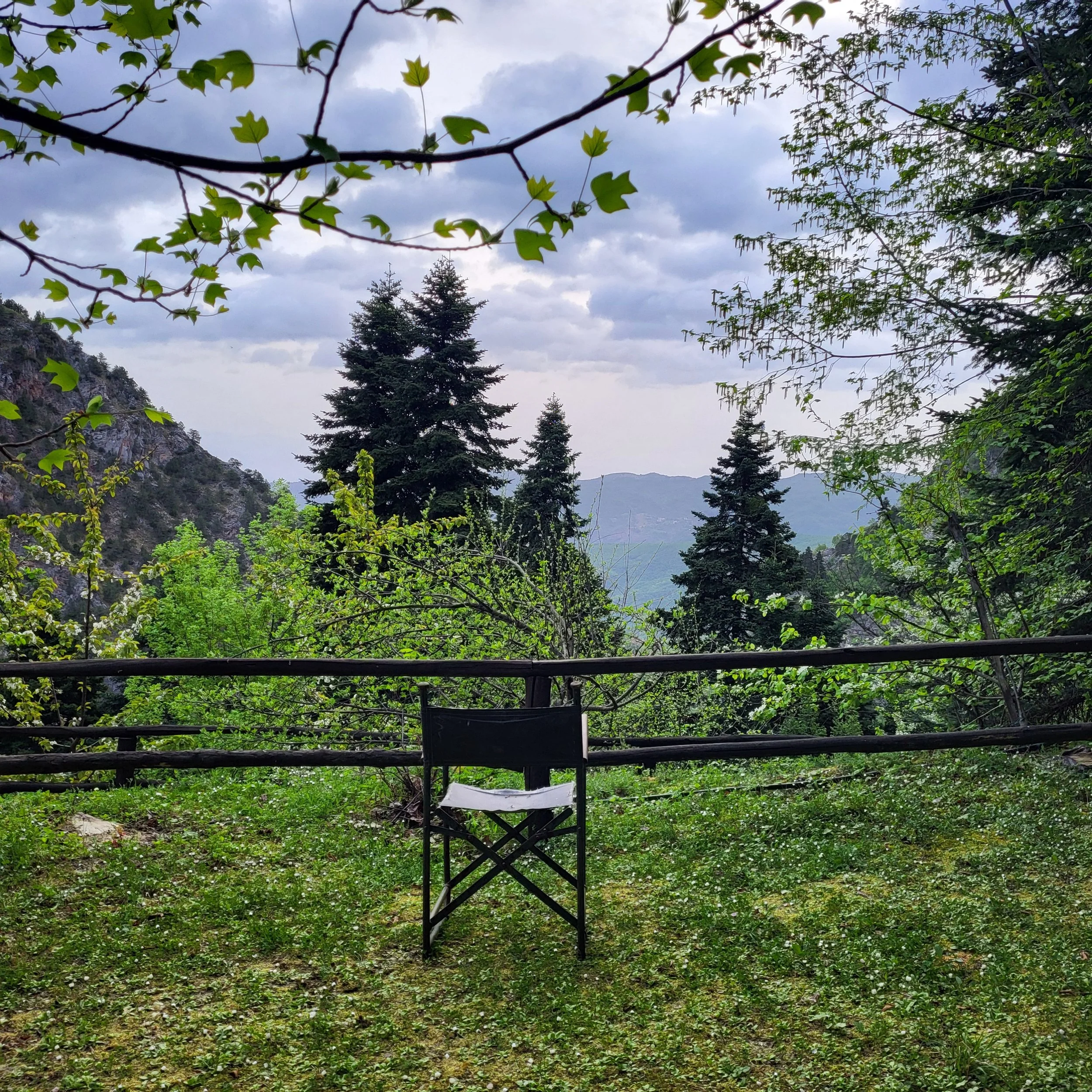 Une chaise pliante noire sur une pelouse verte, avec une barrière en bois devant un paysage de montagnes, de conifères et de ciel nuageux, entourée d'arbres et de feuillage en premier plan.