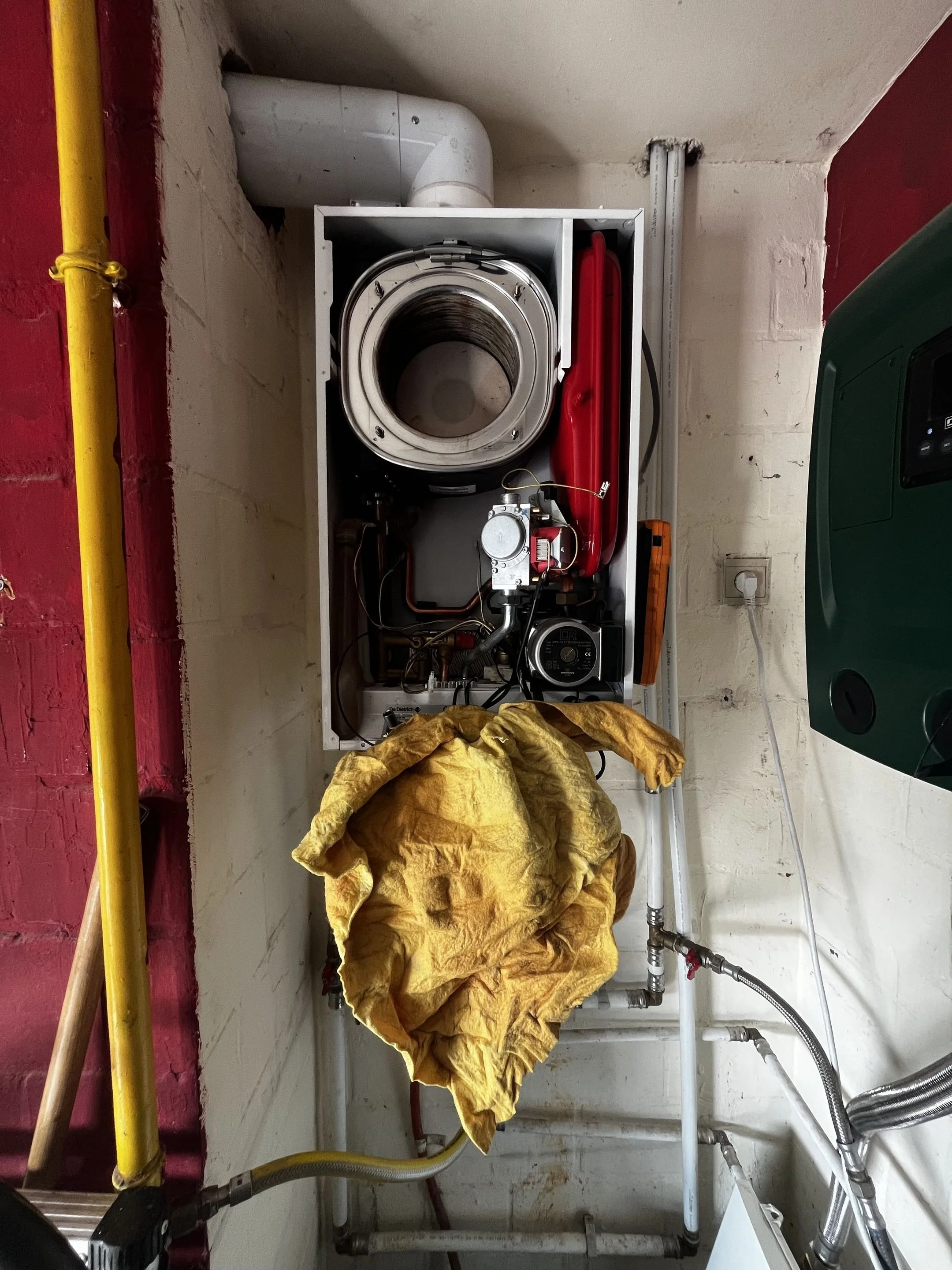 An industrial machine with a large metal barrel, red components, and various pipes and wiring, with a yellow rag draped over part of it, set against a white and red brick wall.