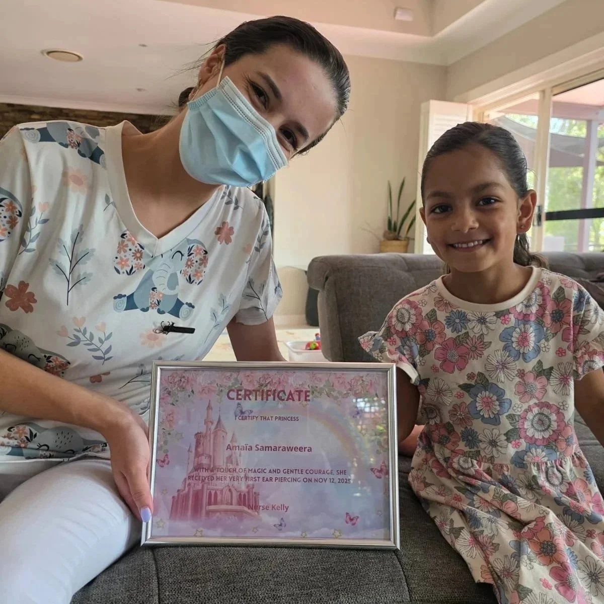 A young girl with dark hair and a floral dress sits on a couch next to a woman with dark hair, a face mask, and a colorful elephant print shirt. The woman holds a framed certificate for a first ear piercing, awarded on November 12, 2025.