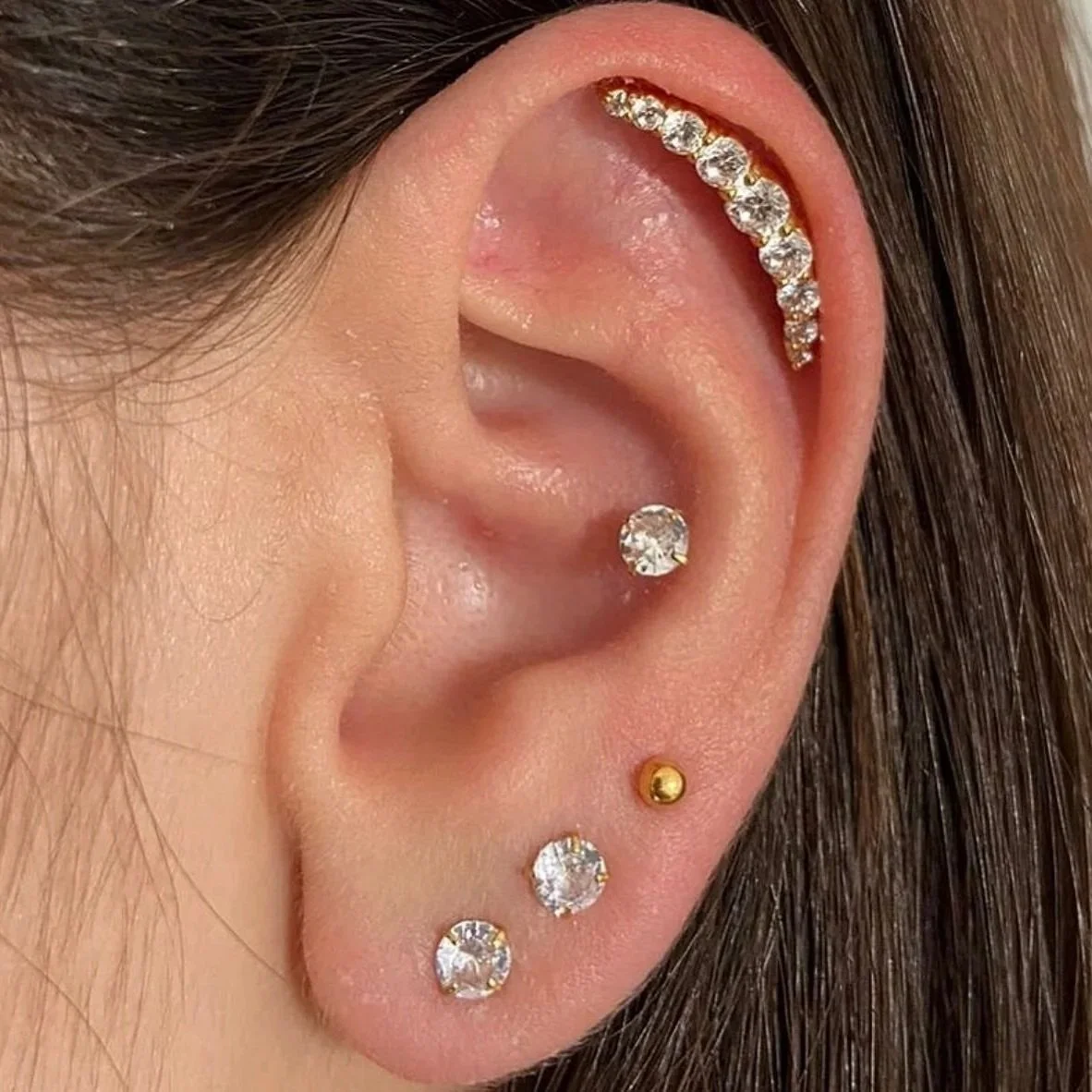 Close-up of a person's ear with multiple piercings, including a gold and diamond stud, two clear gemstone studs, a gold ball stud, and a curved row of diamonds along the upper cartilage.