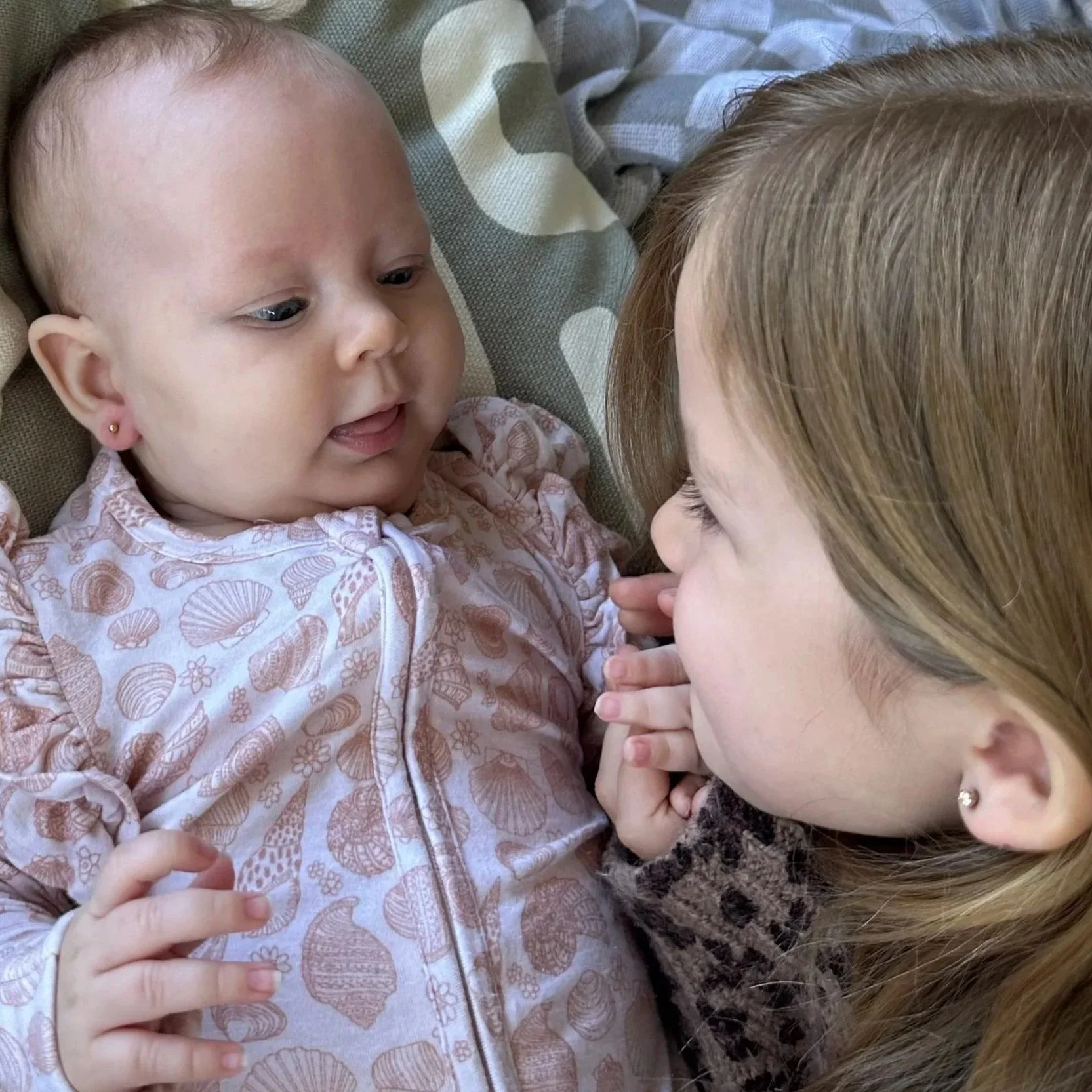 A young girl and a baby girl are lying down facing each other, touching noses and hands, sharing a tender moment.