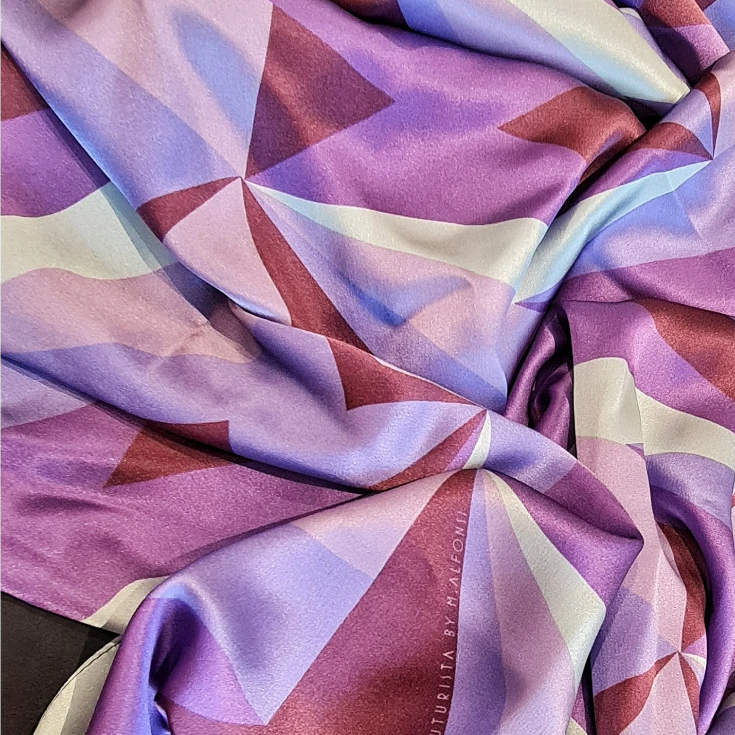 Close-up of a silk scarf with a geometric pattern in shades of purple, pink, and white, featuring triangular shapes and gradient colors.