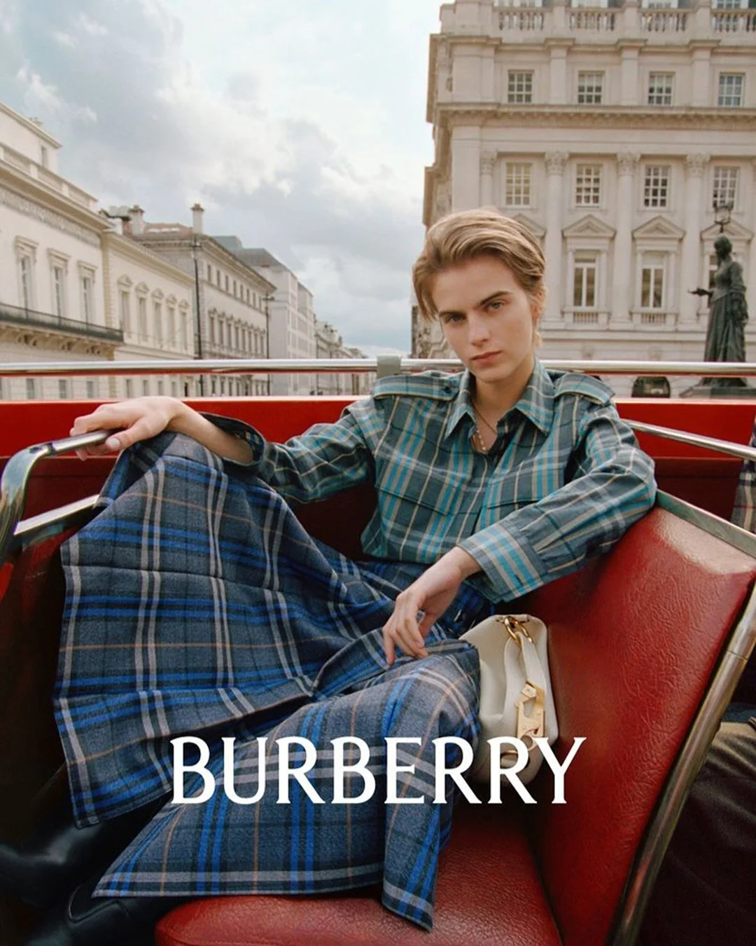 A young person with short hair sitting on the red seats of an open-double-decker tour bus, wearing a green plaid shirt and matching plaid pants, with a cityscape of classical European buildings in the background, and the word 'BURBERRY' overlaid on t