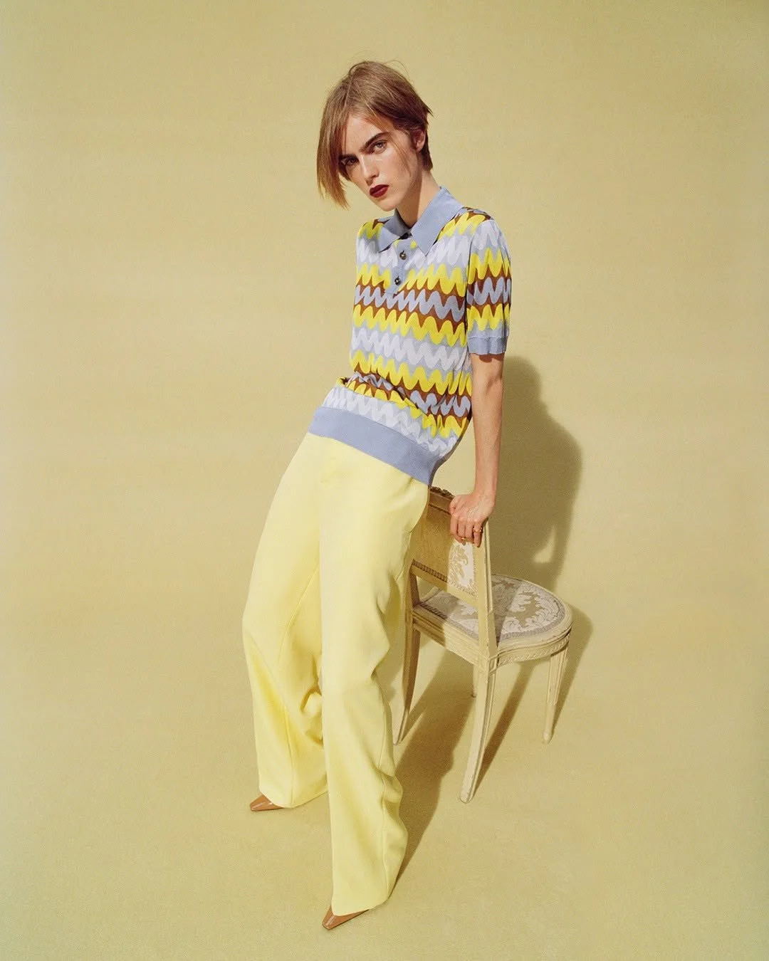 A young woman with short, reddish-brown hair and bold makeup, wearing a multicolored zigzag pattern top and high-waisted yellow trousers, leaning on a white vintage chair with a floral seat cushion against a plain light yellow background.