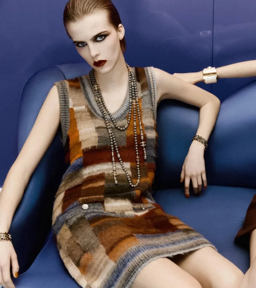 A fashion model with striking makeup and accessorized with multiple necklaces and bracelets, sitting on a blue couch against a blue background, wearing a sleeveless, multicolored plaid dress.