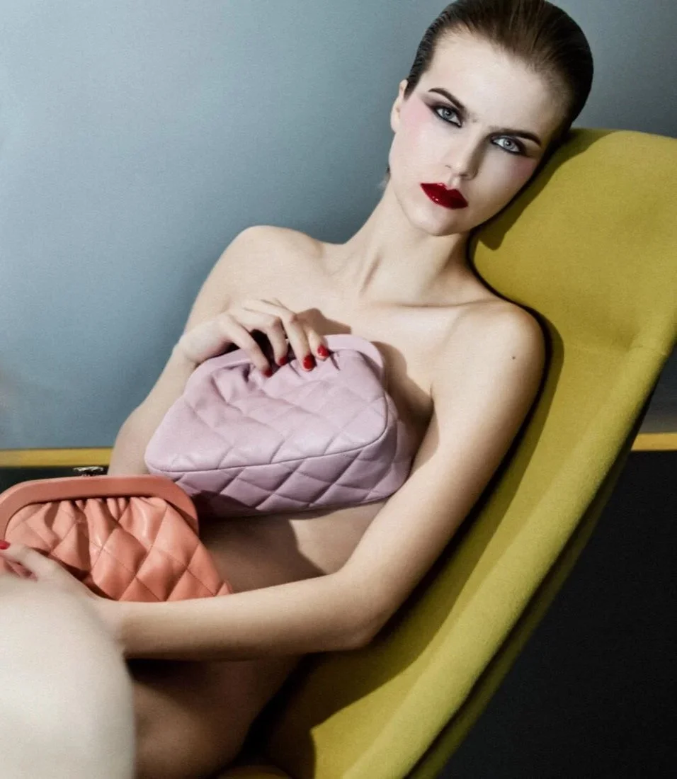 A woman with dark hair and makeup, wearing red lipstick and nail polish, sits nude on a yellow lounge chair, holding two pink quilted handbags.