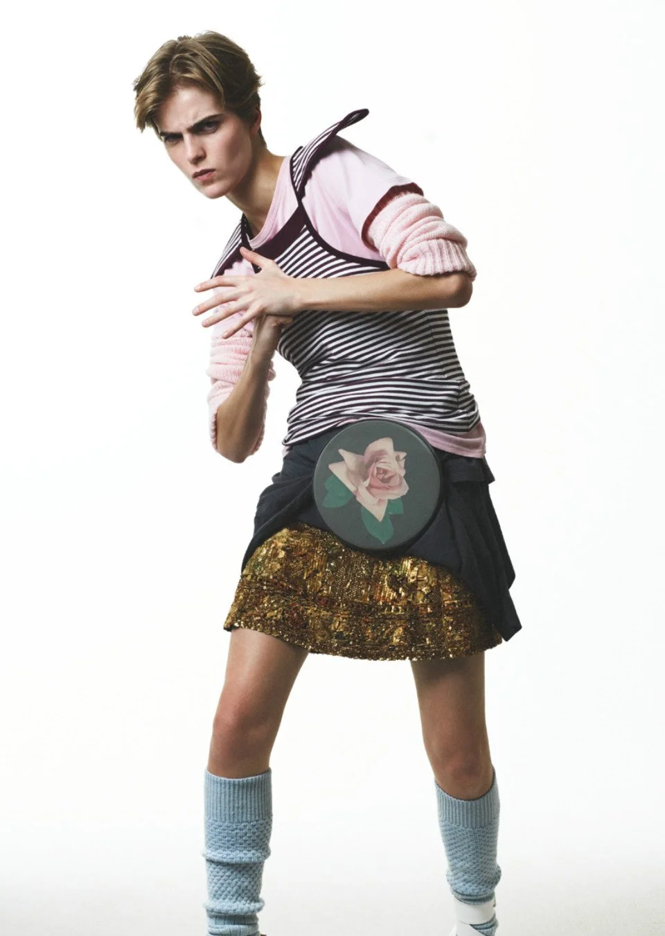 Young woman in trendy clothing with a bold expression, wearing a striped top, metallic gold skirt, and leg warmers, posing against a plain white background.