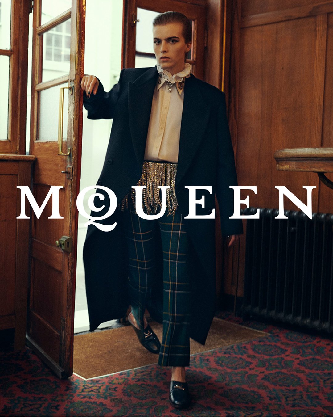 Fashionably dressed person in a black blazer, beige blouse with ruffled collar, pinstriped checkered trousers, and black shoes exits from a wooden door in a room with wooden walls and a red patterned carpet.
