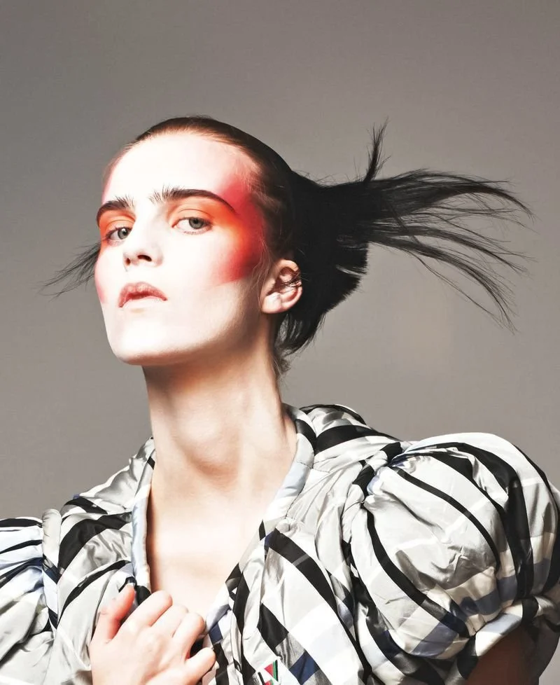 A fashion model with striking makeup and a stylish black-and-white patterned outfit posing against a neutral background.