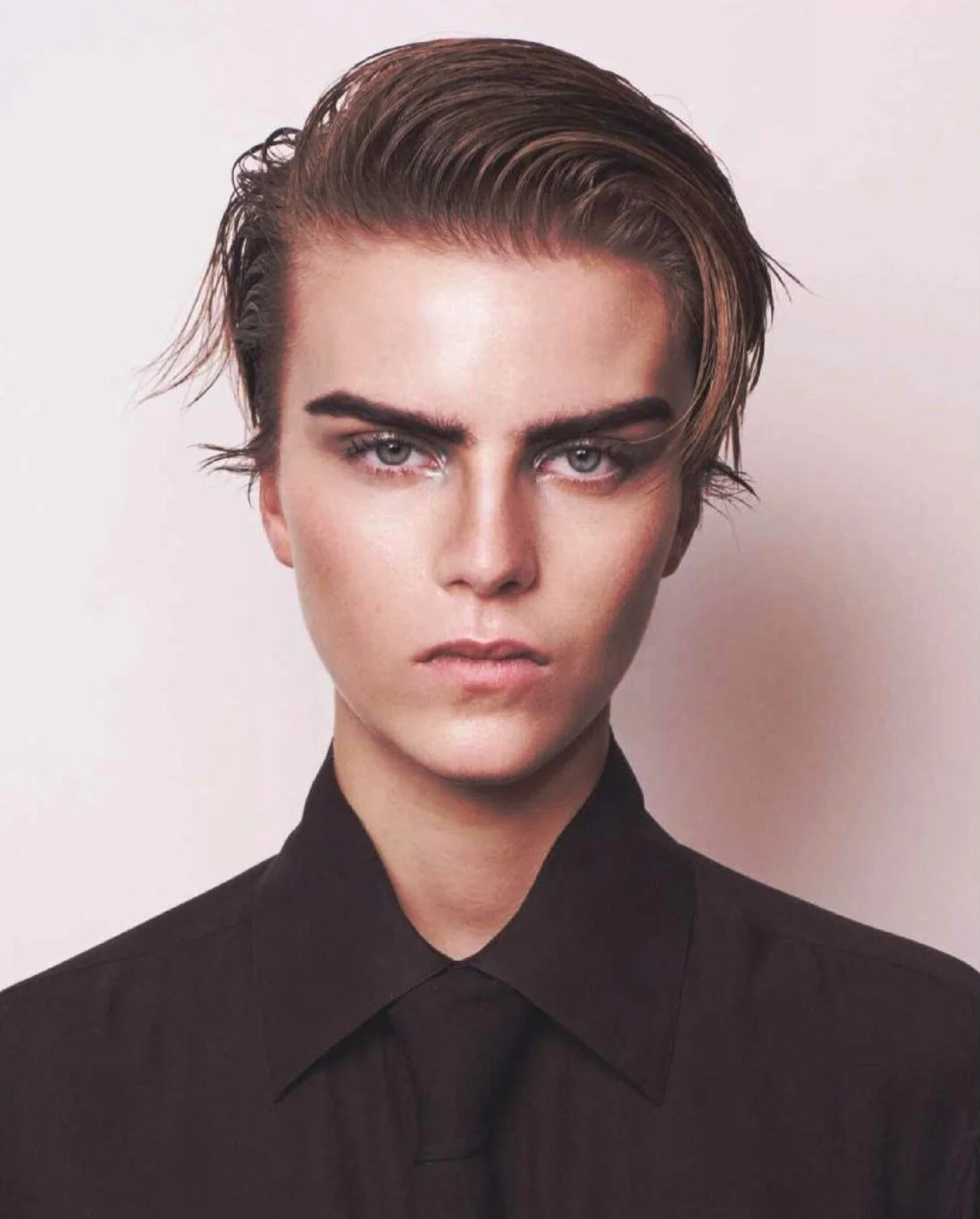 A young person with short, slicked-back hair, wearing a black collared shirt, and with intense blue eyes staring directly at the camera.