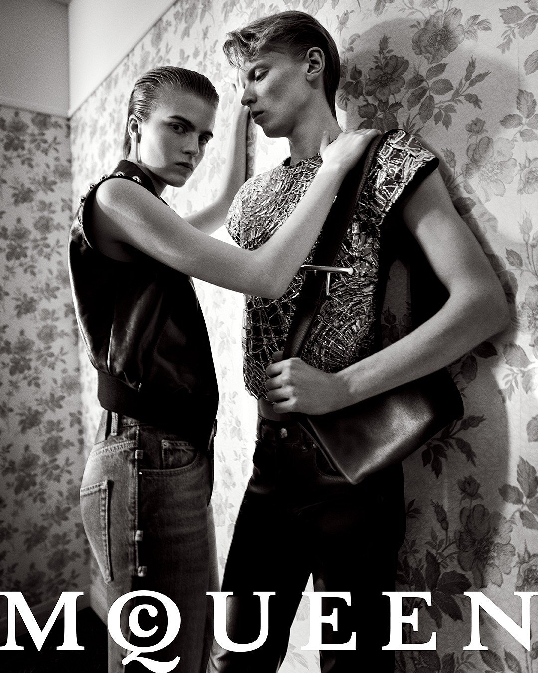 Black and white fashion photo of a male and female models posing against a floral wallpaper background, wearing stylish clothing.