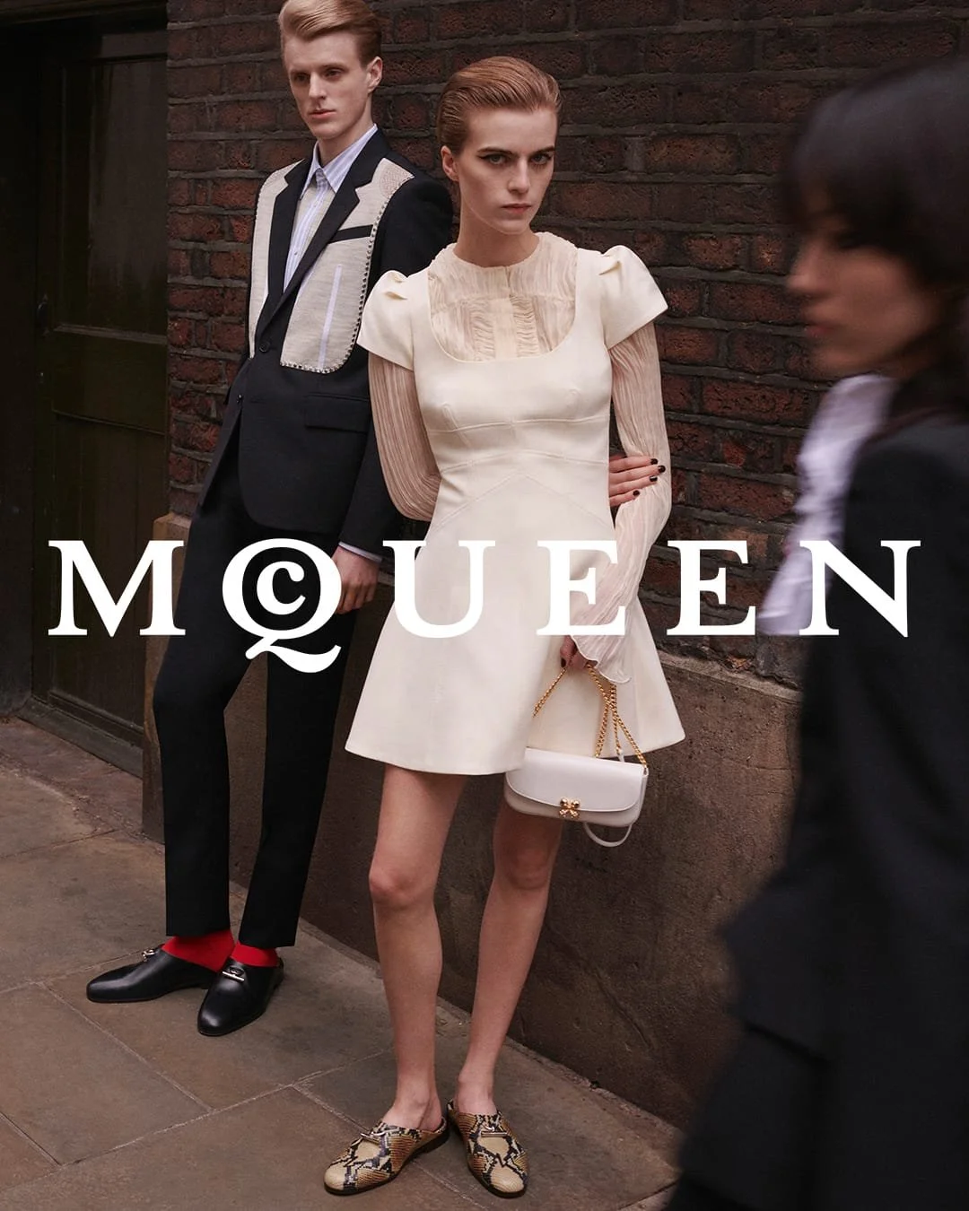 Fashion advertisement featuring three models standing against a brick wall. The woman in the front wears a cream-colored dress, holding a white handbag with gold chain. The man behind her wears a black suit with white and beige details, and the woman