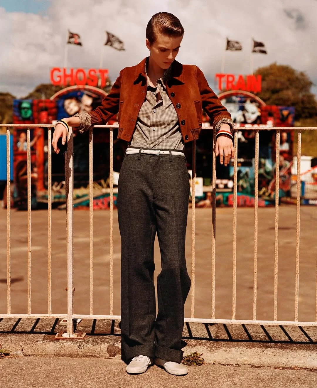 A person stands behind a white metal fence at a fairground, wearing a brown leather jacket, checkered shirt, dark pants, and white shoes, with an amusement park ride in the background and a sign that reads 'GHOST TRAIN'.
