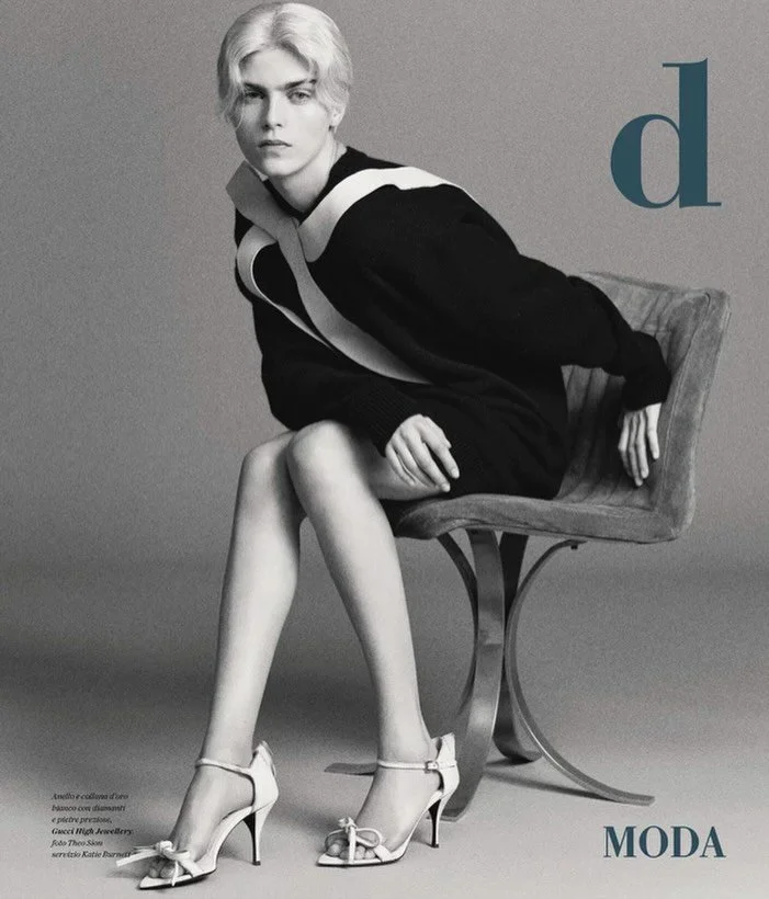 Black and white fashion photograph of a woman sitting on a chair, wearing high heels and a stylish black dress with white accents, with a large blue letter 'd' and the word 'MODA' in the background.