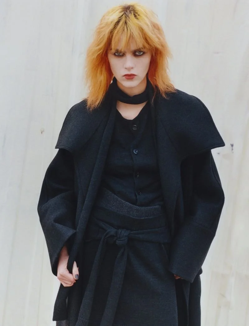 Woman with orange hair wearing dark clothing and a black scarf, standing against a light wooden wall.