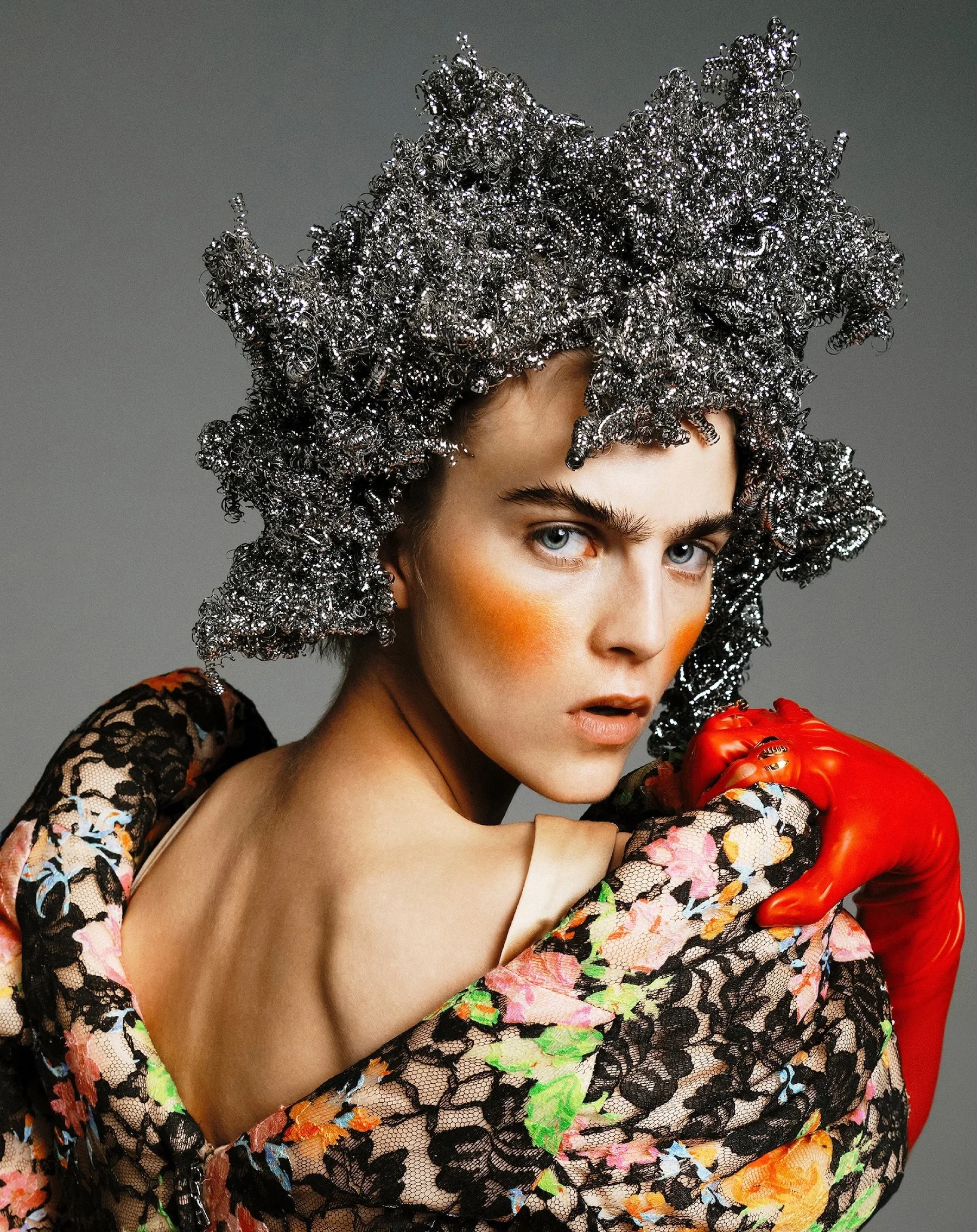 A woman with blue eyes and bold makeup wears a metallic curly headpiece, black lace floral-patterned garment, and bright red gloves, holding a red pepper against a gray background.
