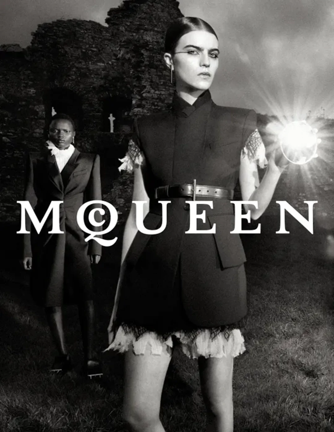 A black and white fashion advertisement featuring two women dressed in high-fashion outfits. The woman in the foreground has dark hair, bold eyebrows, and is holding a mirror or reflective object blinding the camera. The woman in the background is we