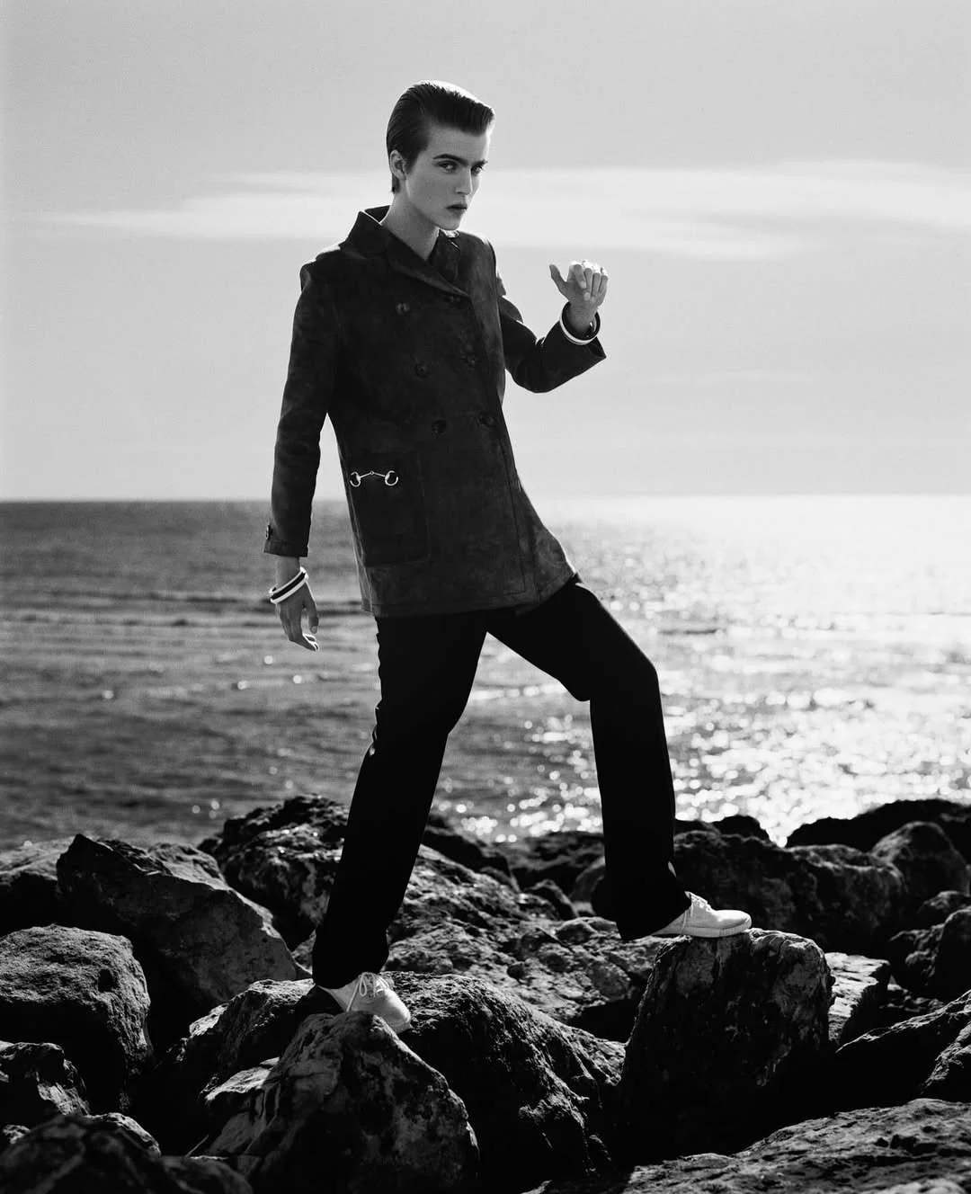 Black and white photo of a woman standing on rocks by the sea, wearing a raincoat and walking shoes, with a confident pose and a serious expression.