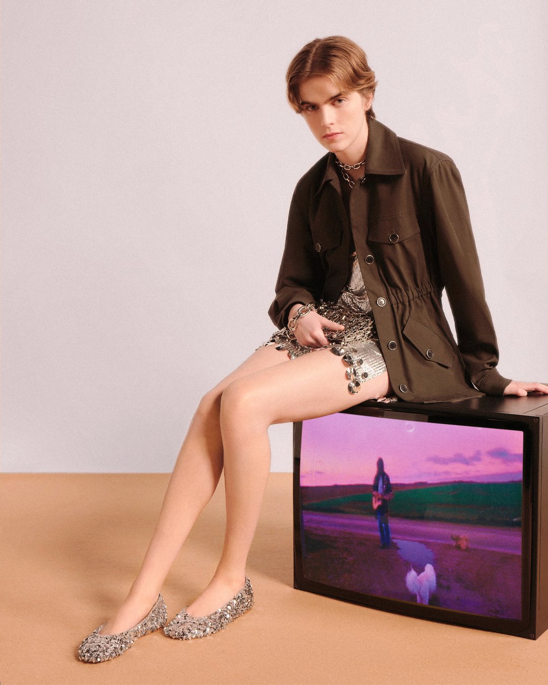 Young person sitting on a black TV with a landscape screen; wearing a brown jacket, shiny patterned shorts, and silver glitter shoes.