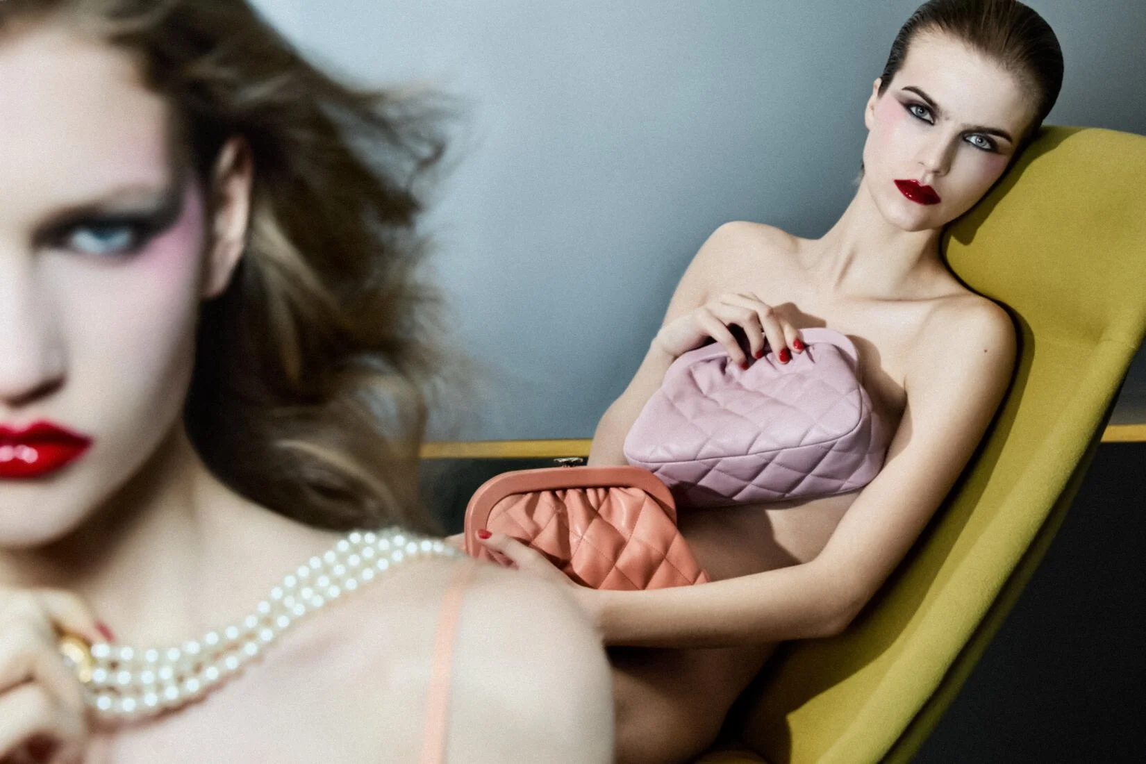 Two women with styled hair and makeup, seated in a modern indoor setting. One woman in the foreground is partially visible, wearing a pearl necklace with bold red lipstick. The second woman is sitting on a yellow chair, holding pink and orange quilte