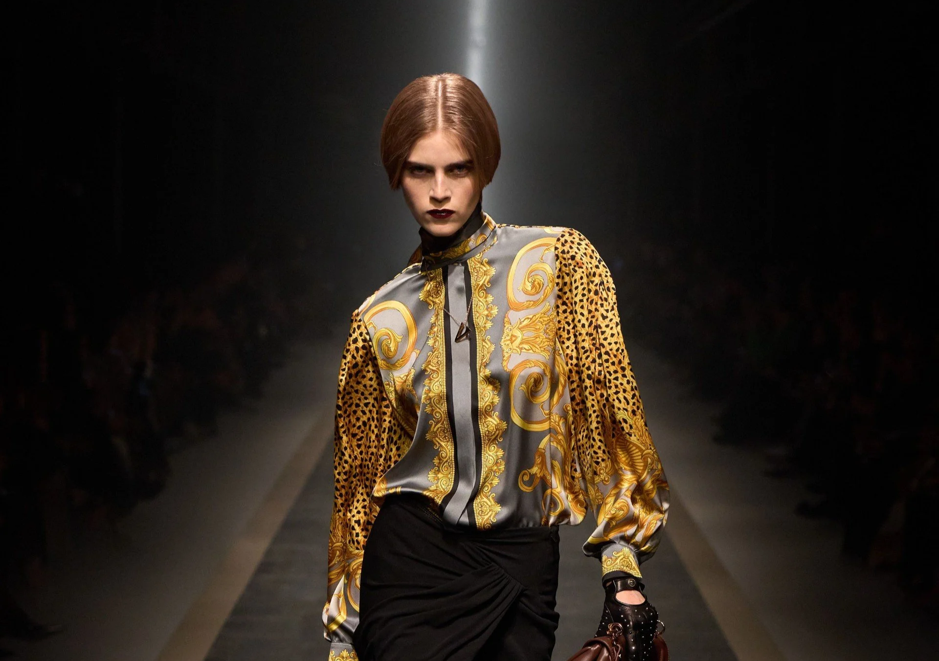 A female model walking on a runway during a fashion show, wearing a ornate patterned blouse with gold and silver tones, black skirt, and black footwear, with a dark background and spotlight overhead.