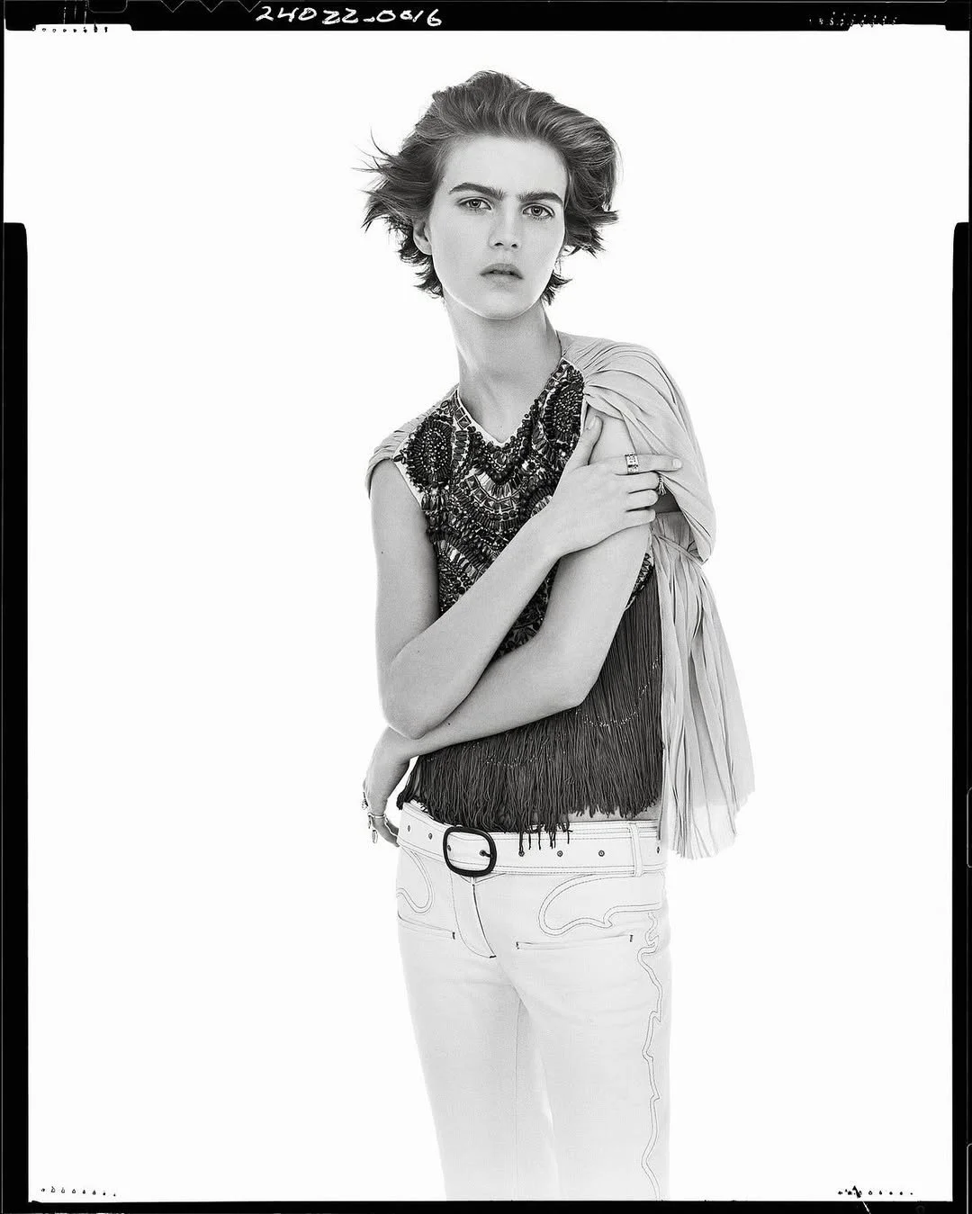Black and white studio portrait of a woman with short wavy hair, wearing a sleeveless embroidered top, light-colored pants, and multiple rings, with her arms crossed and holding her shoulder.