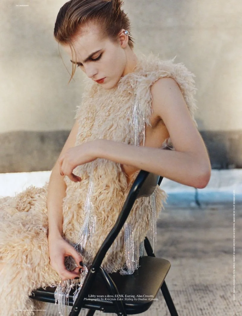 A young woman with styled hair and makeup wearing a textured beige dress with silver fringe accents, sitting on a black chair.