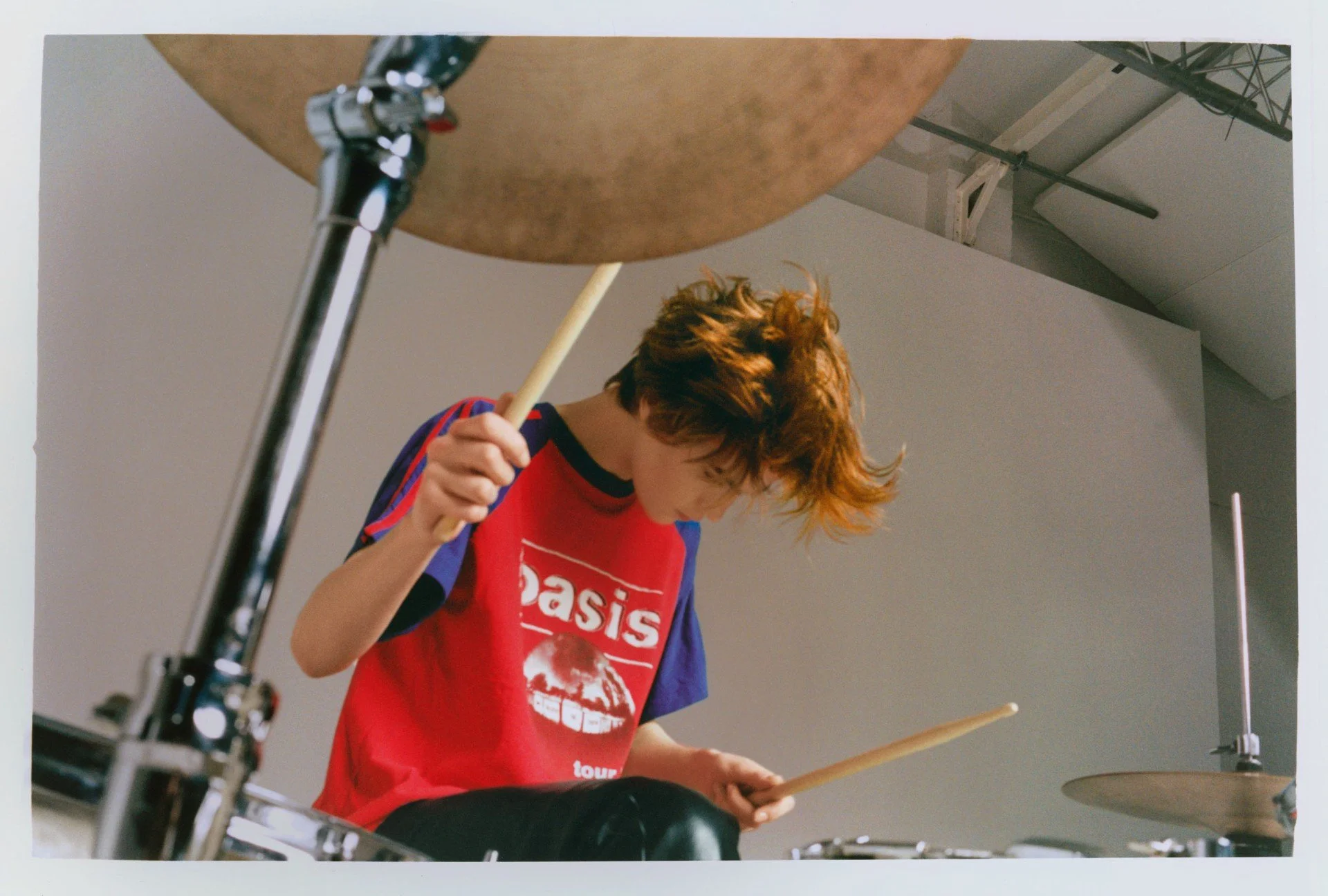 Person with curly hair playing drums in a studio or rehearsal space, wearing a red Oasis tour T-shirt.