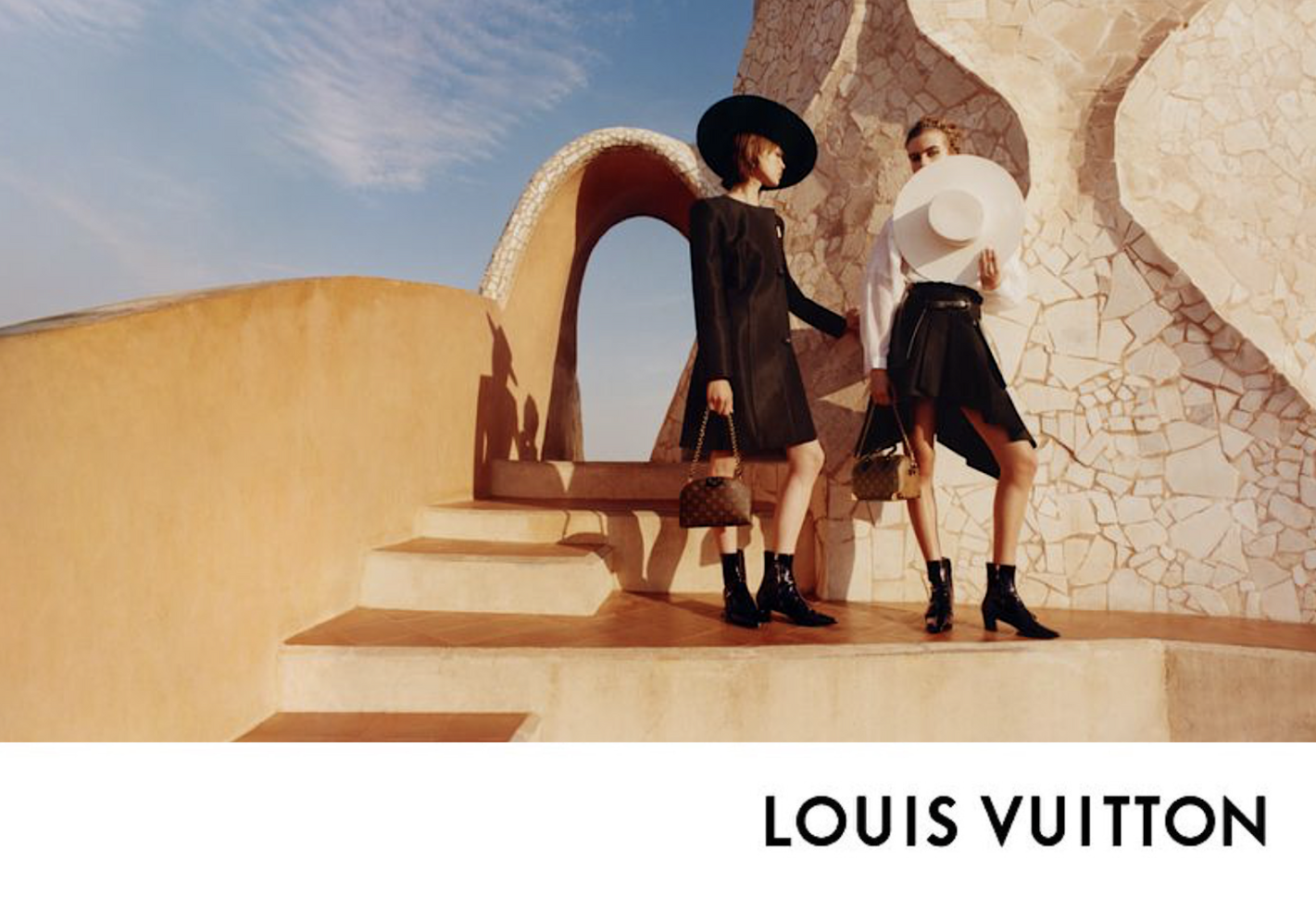 Two women wearing fashionable black and white outfits with large hats, holding designer handbags, standing on steps against an architectural backdrop with a curved wall and sky.