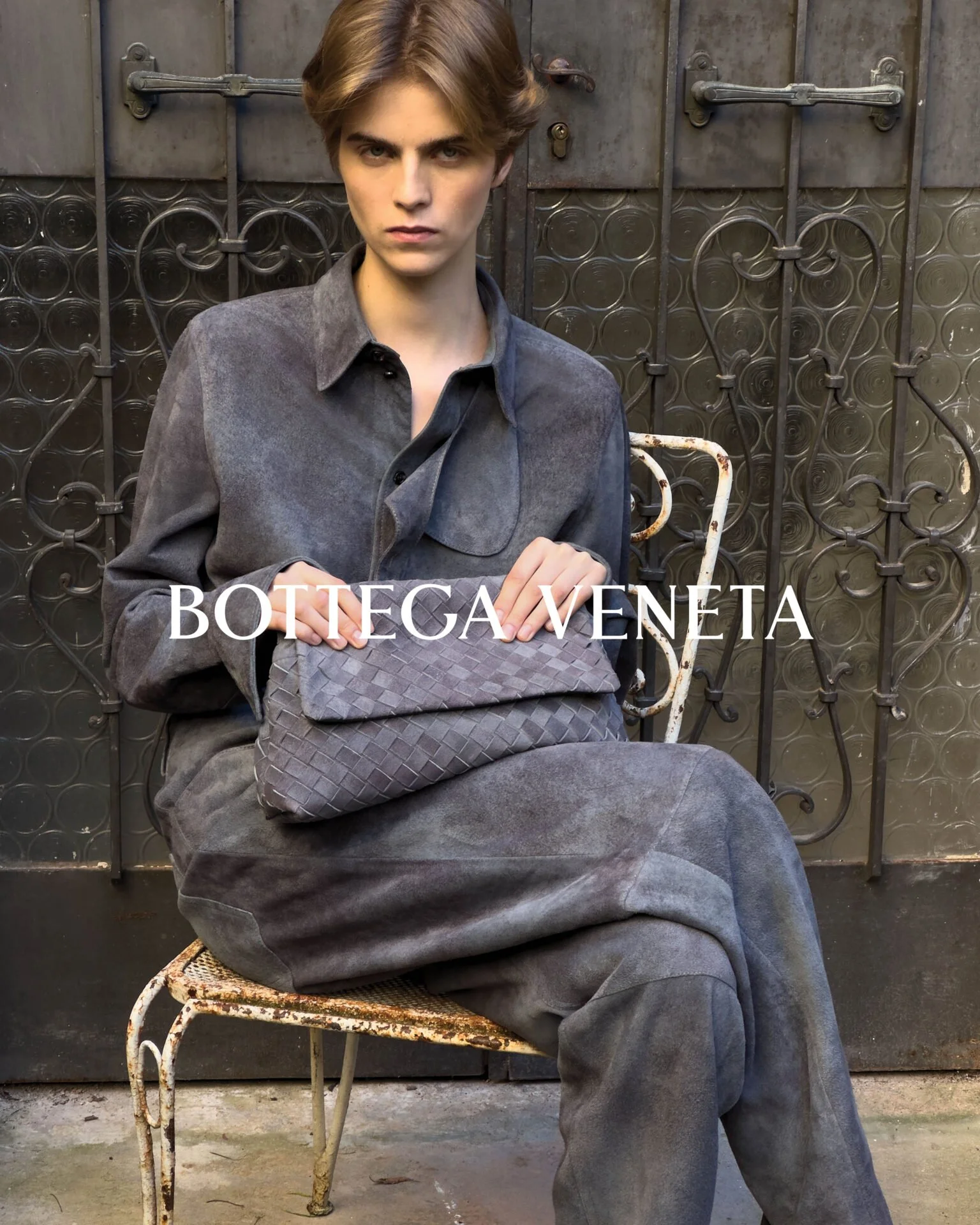 A young man with short brown hair sitting on a rusted white metal chair in front of a metal door with decorative ironwork. He is wearing a gray suede shirt and gray pants, with a serious expression, hands resting on his lap, and a woven gray bag on h