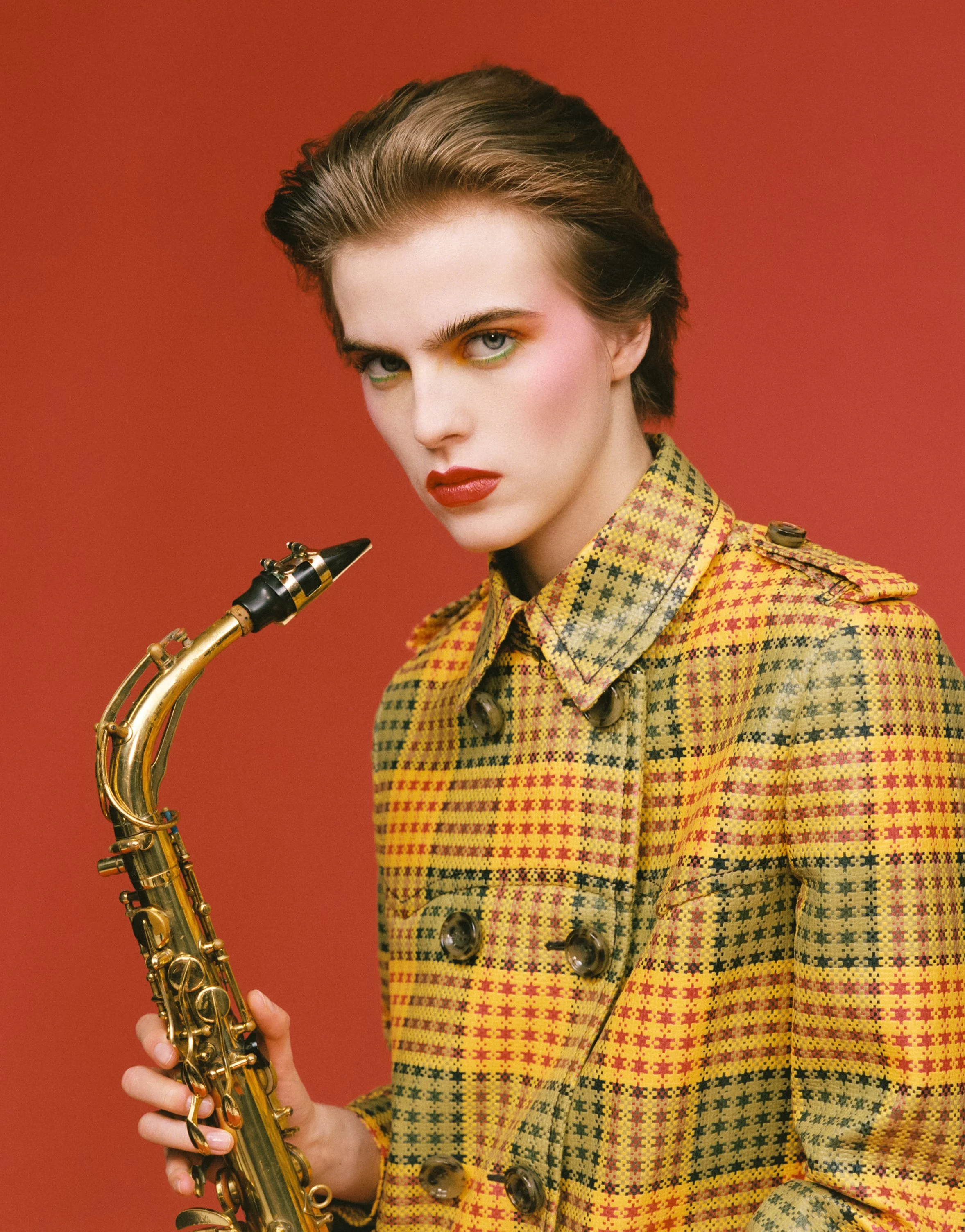 Close-up of a person holding a saxophone against a red background, wearing a yellow and red patterned jacket, with short styled brown hair, bold makeup, emphasizing their face and expression.