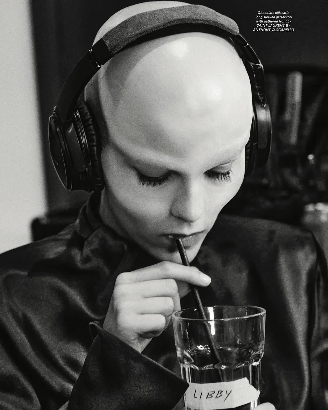 A black and white photo of a young woman with a bald head, wearing large over-ear headphones and a leather jacket, sipping a drink through a straw in a glass labeled 'Libby'.