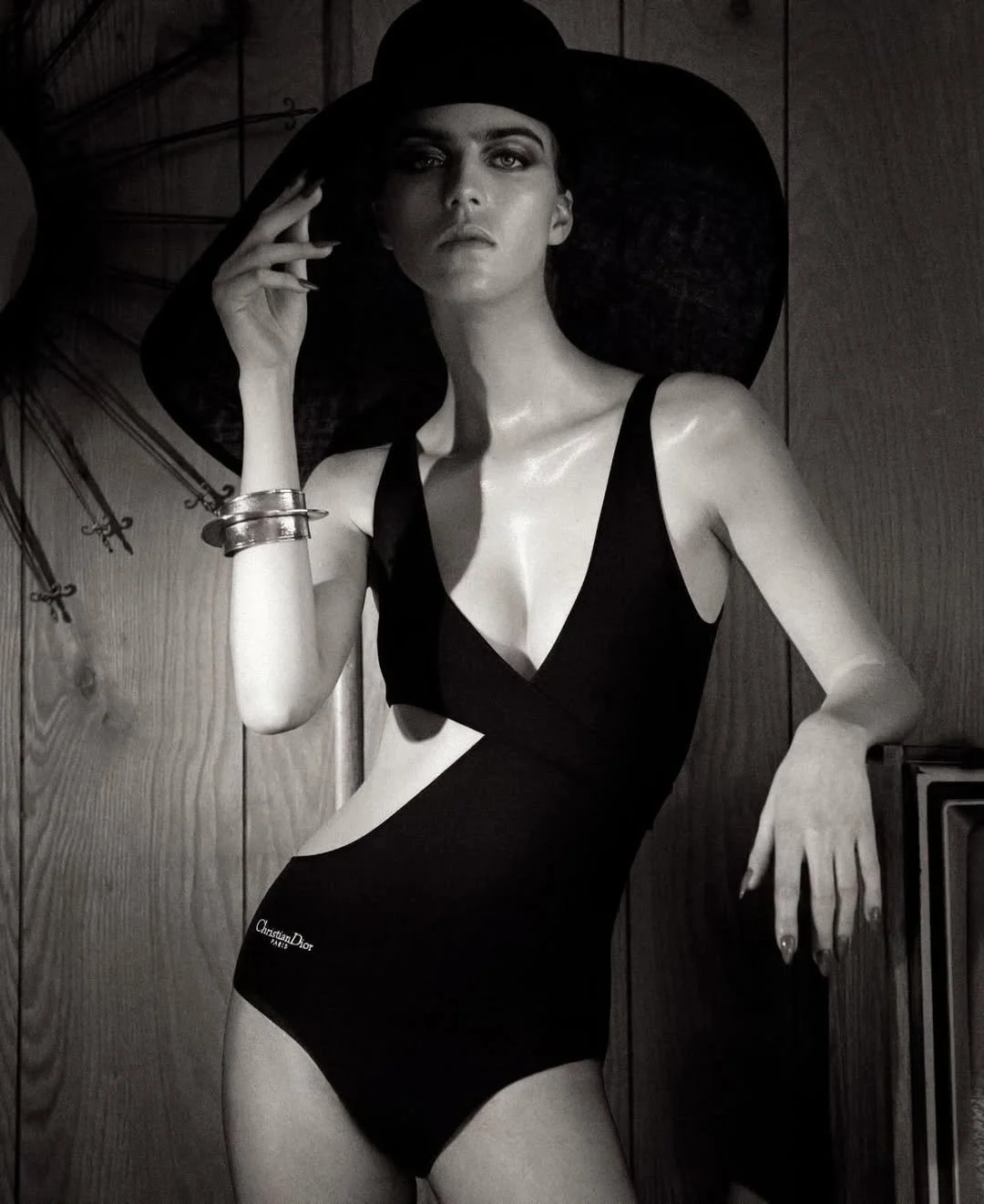 Black and white photo of a woman wearing a black one-piece Christian Dior swimsuit and a wide-brimmed hat, leaning against a wooden wall.