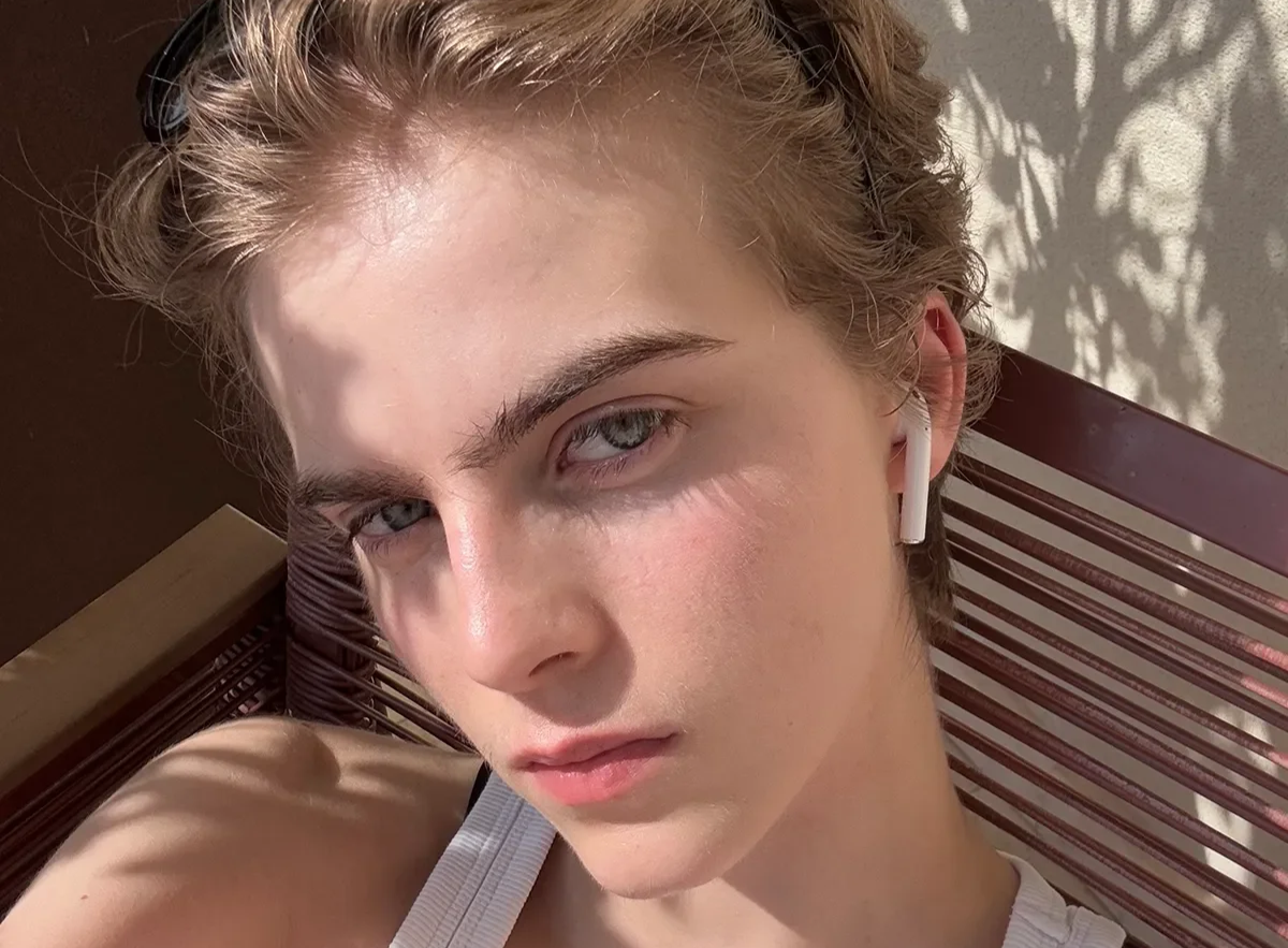 Close-up of a young woman with blonde hair and blue eyes taking a selfie, wearing white earbuds and a white tank top, sitting on a wooden bench with shadow patterns on the wall behind her.