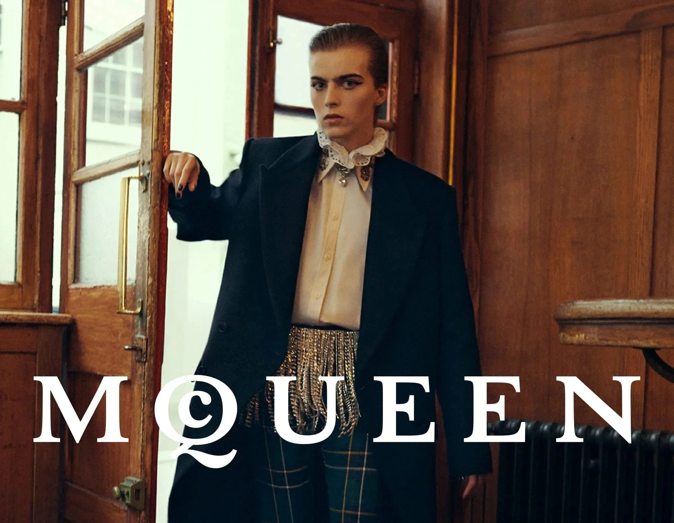 Woman with styled hair and makeup holding a purse, standing in front of wooden doors with a glass window, and a half-visible sign with the partial text 'MOU'.