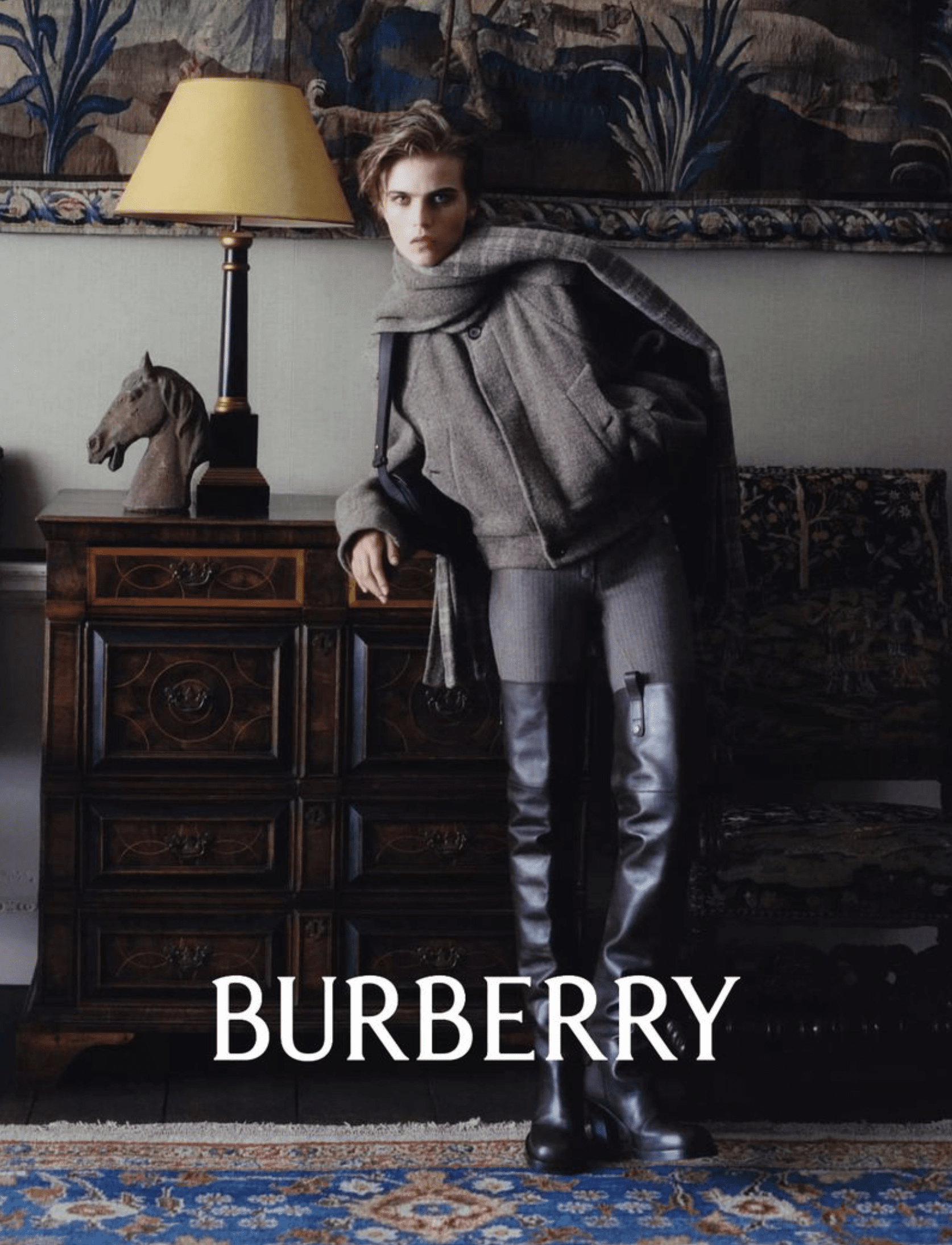 A woman with short hair wearing a gray coat and boots, leaning slightly forward, standing in a room with a large tapestry on the wall, a wooden sideboard with a horse head sculpture, and a lamp with a yellow shade. The word 'BURBERRY' is written at t