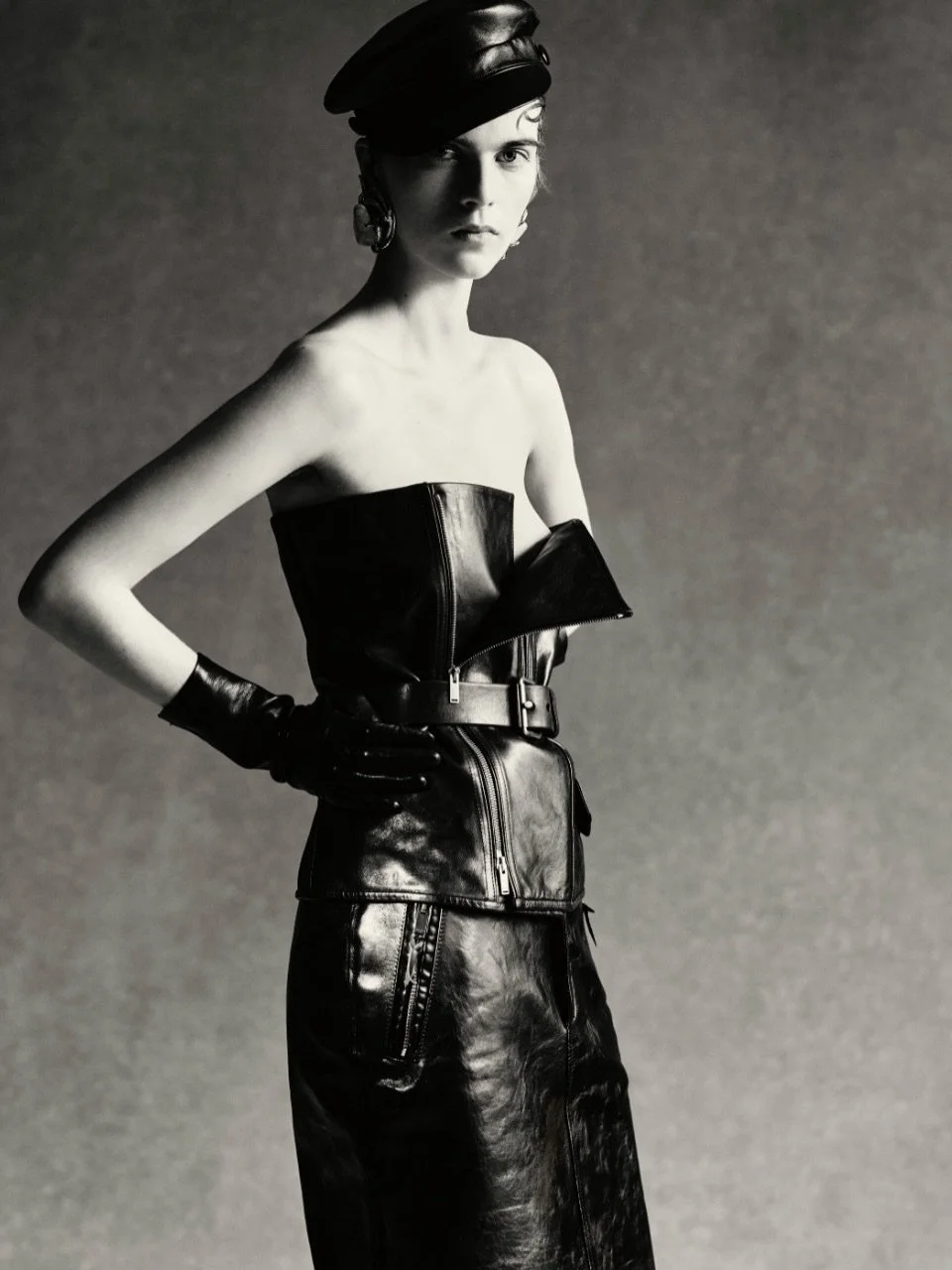 A black-and-white photo of a woman wearing a strapless leather outfit with a zipper, a belt, gloves, and a leather hat. She has earrings and stands with her hand on her hip, looking at the camera.