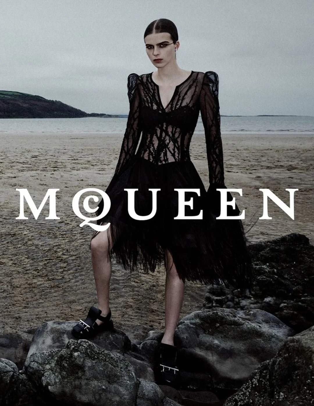 A woman with dark hair and makeup standing on rocks at the beach, wearing a sheer black dress with a fringed skirt, with the word 'MQUEEN' overlaid.