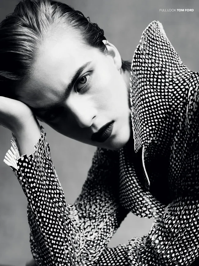 Black and white close-up portrait of a woman with slicked-back hair, wearing a textured patterned jacket, posing with her head resting on her hand, looking directly at the camera.