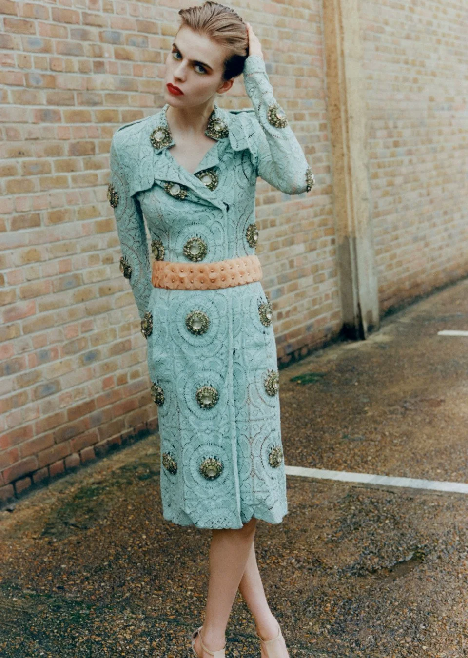 A woman with blonde hair styled back, wearing a vintage-style light blue dress decorated with large ornate embellishments and a peach belt, standing outside against a brick wall.