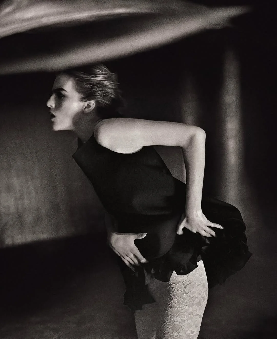 A black and white photo of a woman in a sleeveless black dress, balancing on her right leg with her left hand on her hip and her right hand on her thigh, bending forward with her hair swept back, inside a dark, enclosed space.