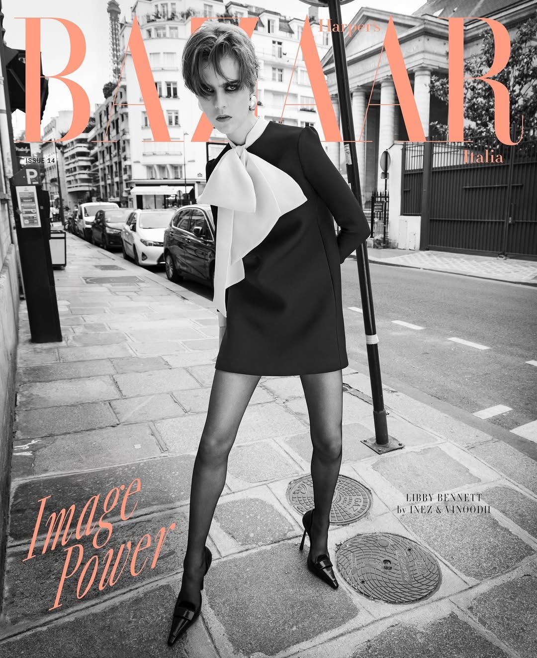 Black and white magazine cover featuring a woman in a black dress with a large white bow, standing on a city sidewalk with parked cars and buildings in the background.