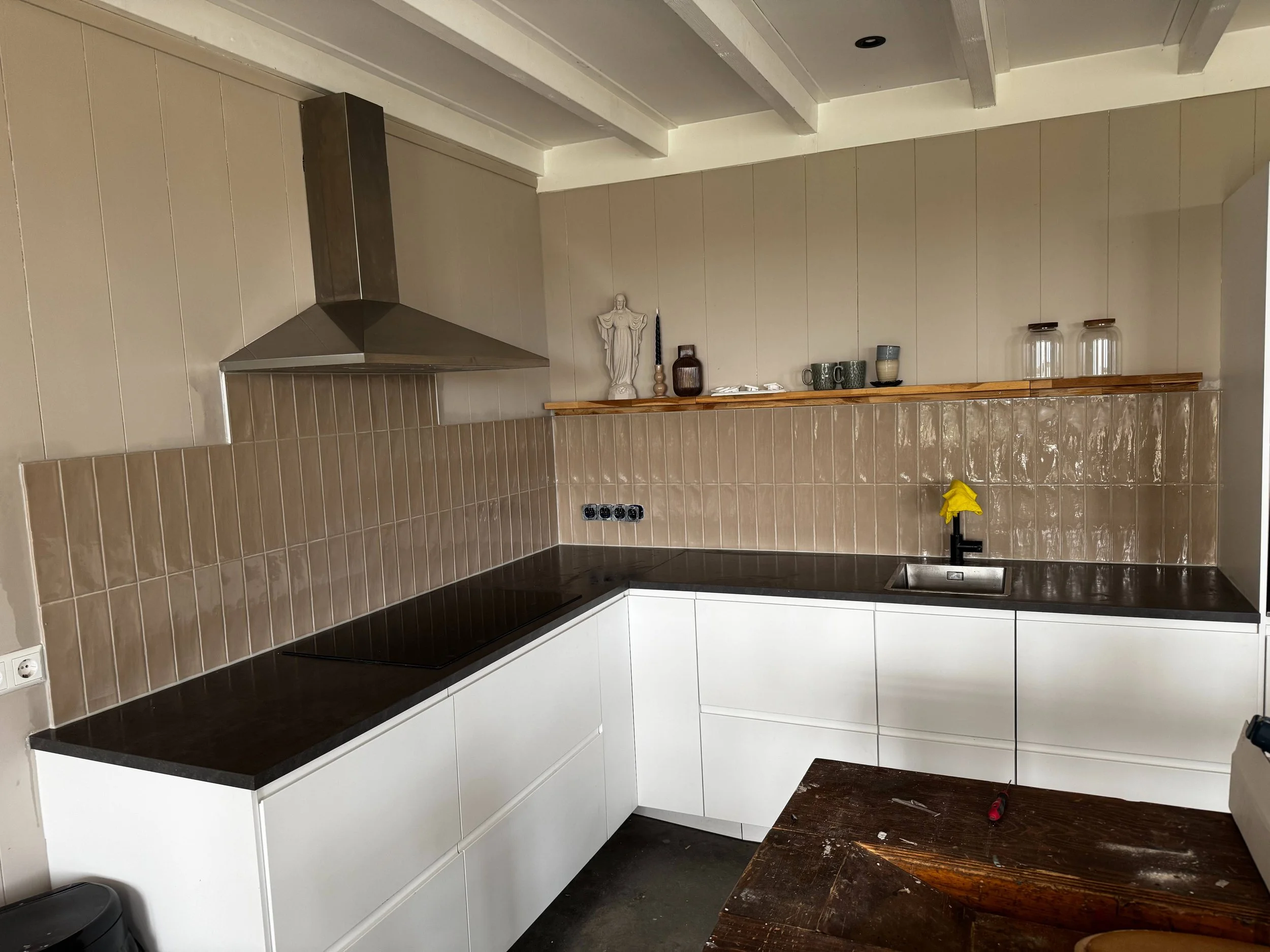 Moderne keuken met zwart aanrecht, witte kasten en beige tegels op de muur, een afzuigkap en open plank met decoratieve items en glazen.