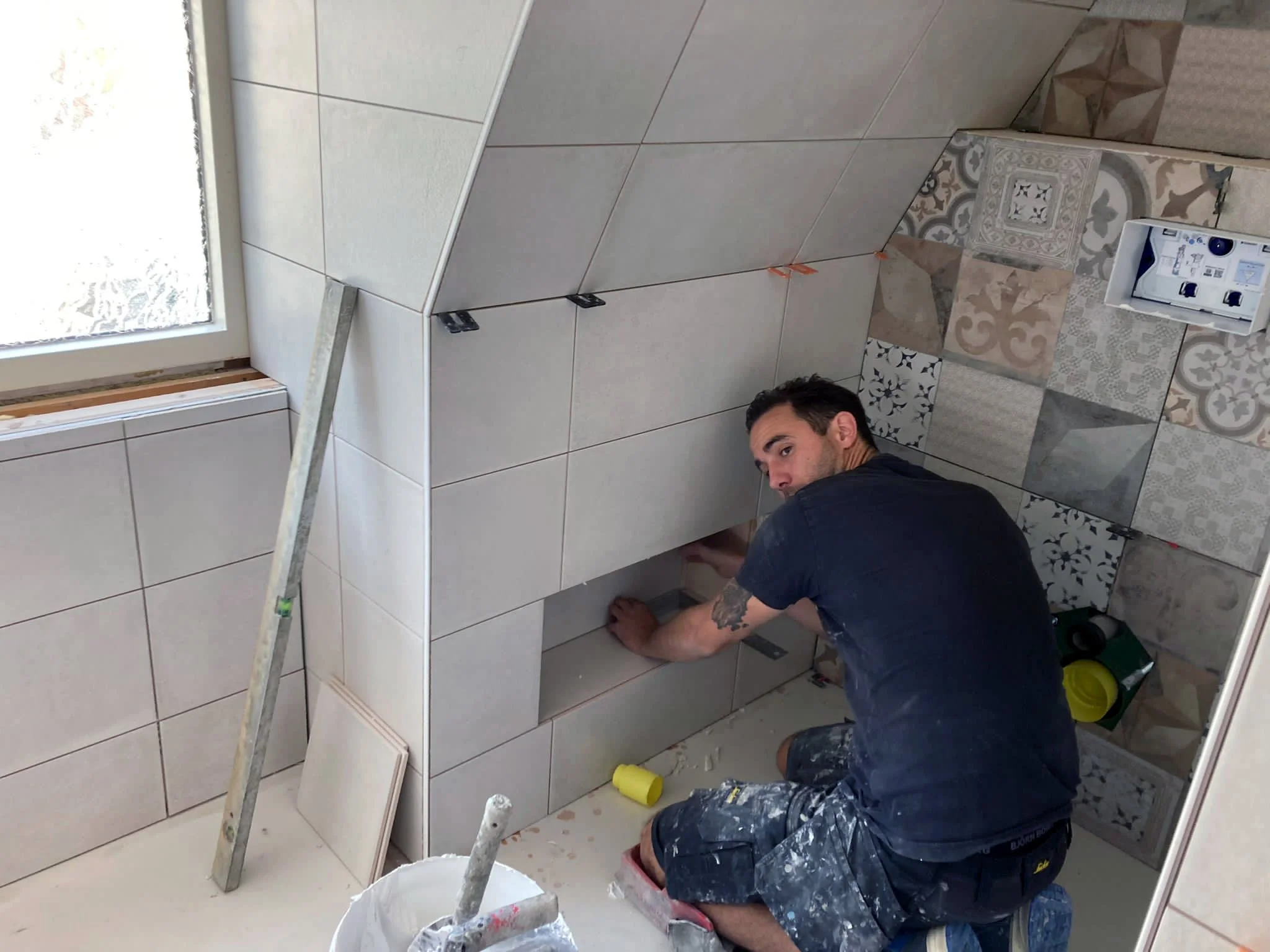 Man die tegels in een kamer plaatst tijdens renovatie, met een ladder en schildermaterialen in de omgeving.